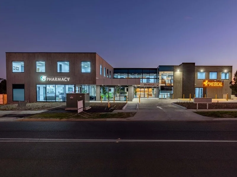 Image of a modern medical facility at dusk, featuring a pharmacy and medical center with illuminated signs and large glass windows.