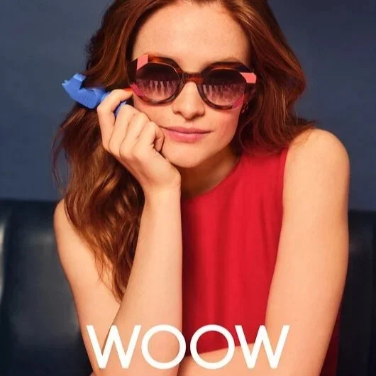A woman with red hair wearing pink WOW sunglasses, available exclusively at Torquay Eyesore and a red sleeveless top, resting her chin on her hand with a playful smile; text overlay says 'WOW'.