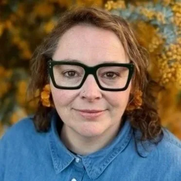 A woman with curly brown hair wearing black glasses and a blue shirt, standing outdoors with yellow autumn leaves in the background.