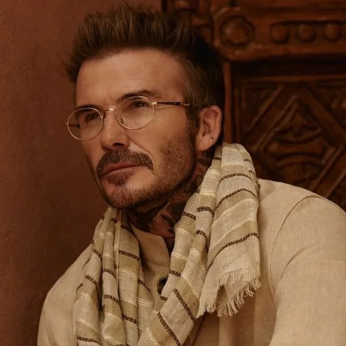 A man wearing beige clothing, glasses, and a striped scarf, sitting in front of a wooden background, with a logo for EYEWEAR by DAVID BECKHAM and branding underneath.