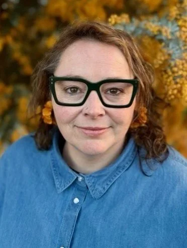A woman with shoulder-length brown hair, wearing black glasses and a blue collared shirt, standing outdoors with yellow flowers in the background.