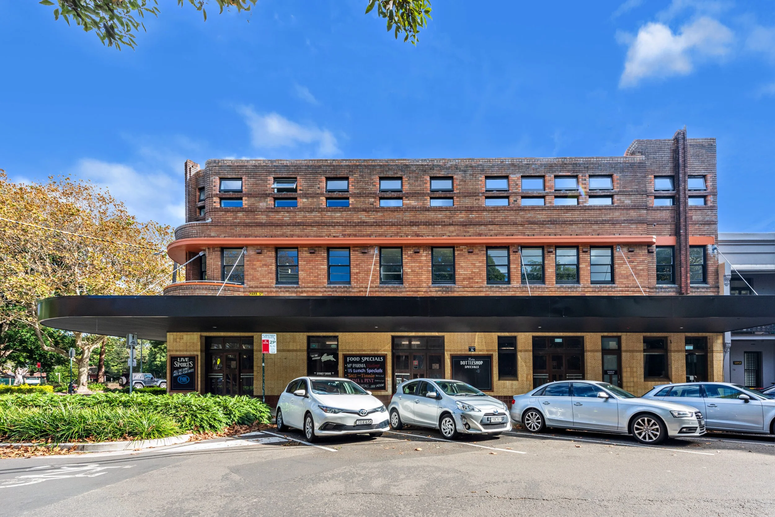 A multi-story brick building with large black overhang on the ground floor, which contains a restaurant with signs displaying food specials, nearby cars parked along the street, and trees with green leaves under a bright blue sky.