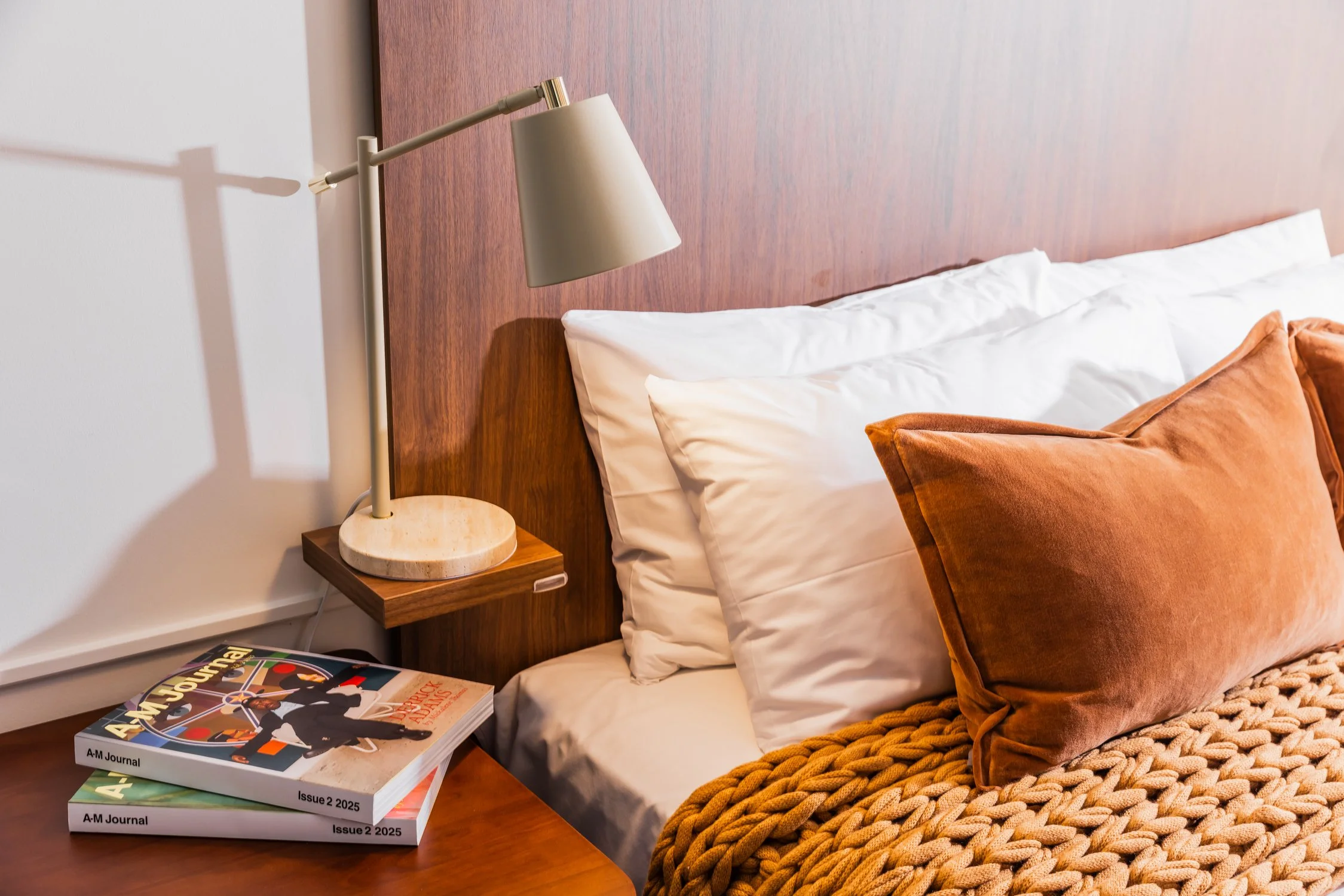 A neatly made bed with white pillows, an orange velvet pillow, and a chunky knit blanket in a cozy bedroom. A modern beige desk lamp, a wooden headboard, and a stack of magazines are on the bedside table.
