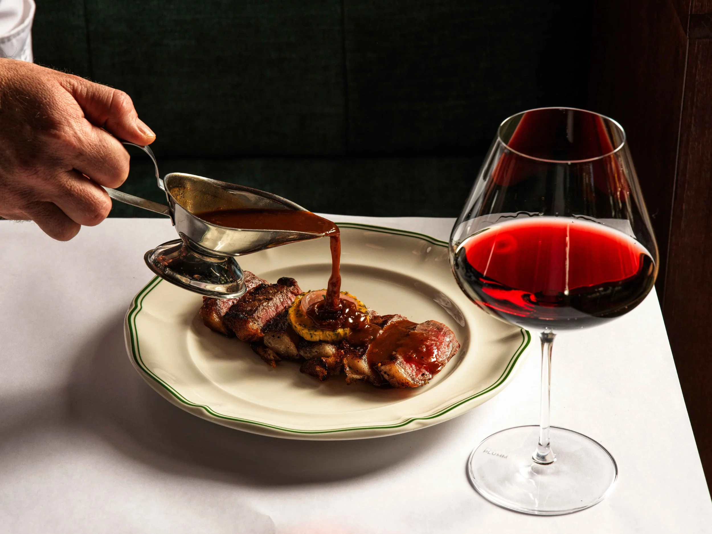 A plate with cooked steak topped with a sauce, a glass of red wine, and a hand holding a gravy boat pouring sauce onto the steak.