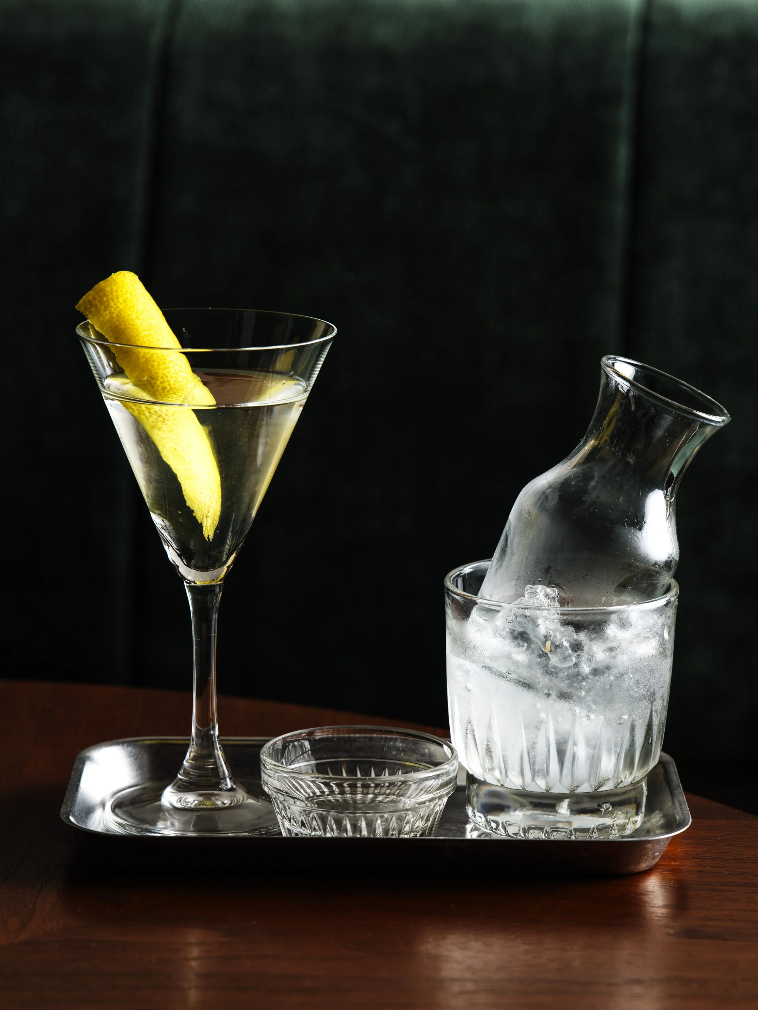 A tray with a tall martini glass containing a lemon wedge and a glass with ice and a carafe on a dark wooden surface.