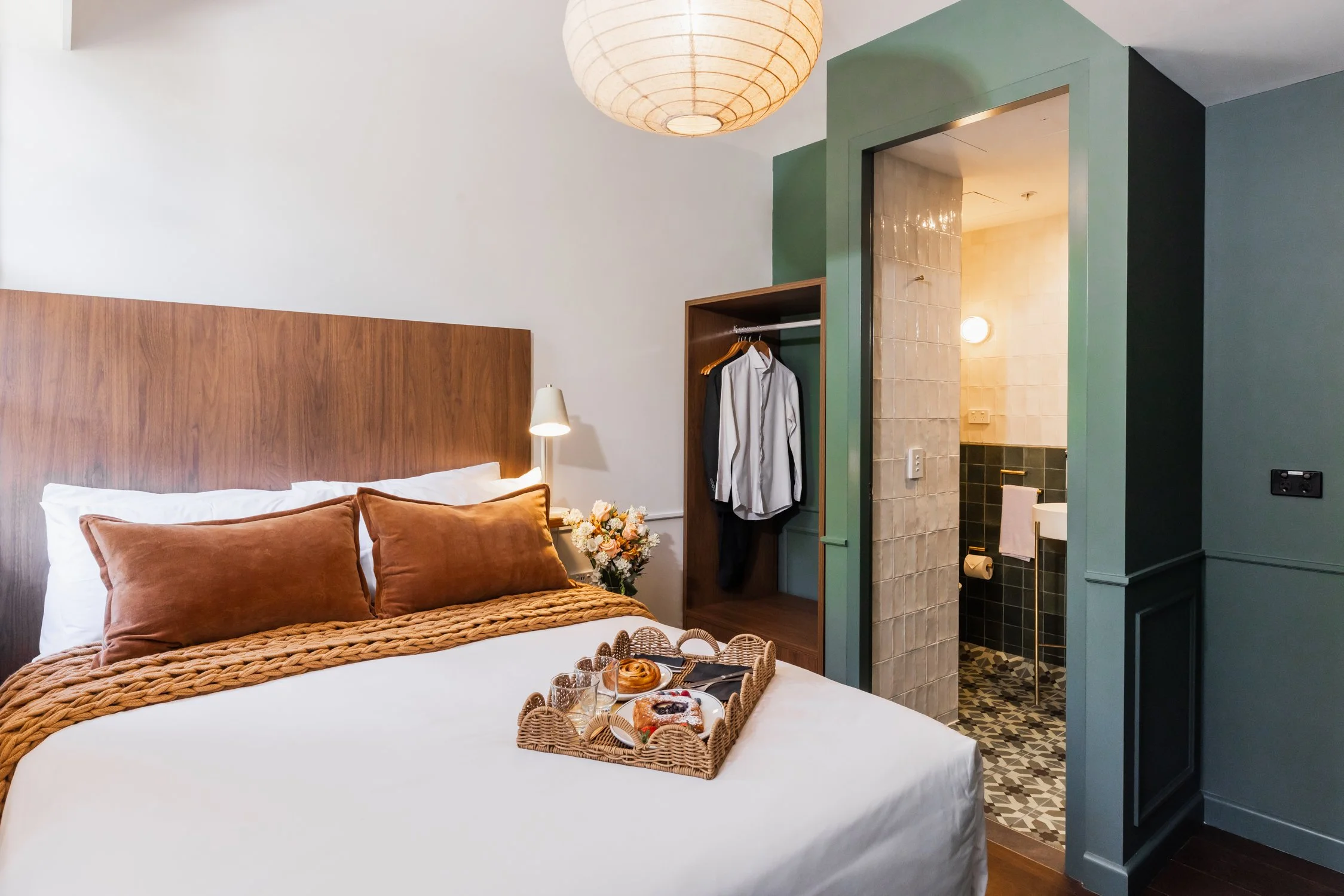 Hotel room with bed, pillows, tray of breakfast items, open wardrobe with clothes, and bathroom visible through doorway.