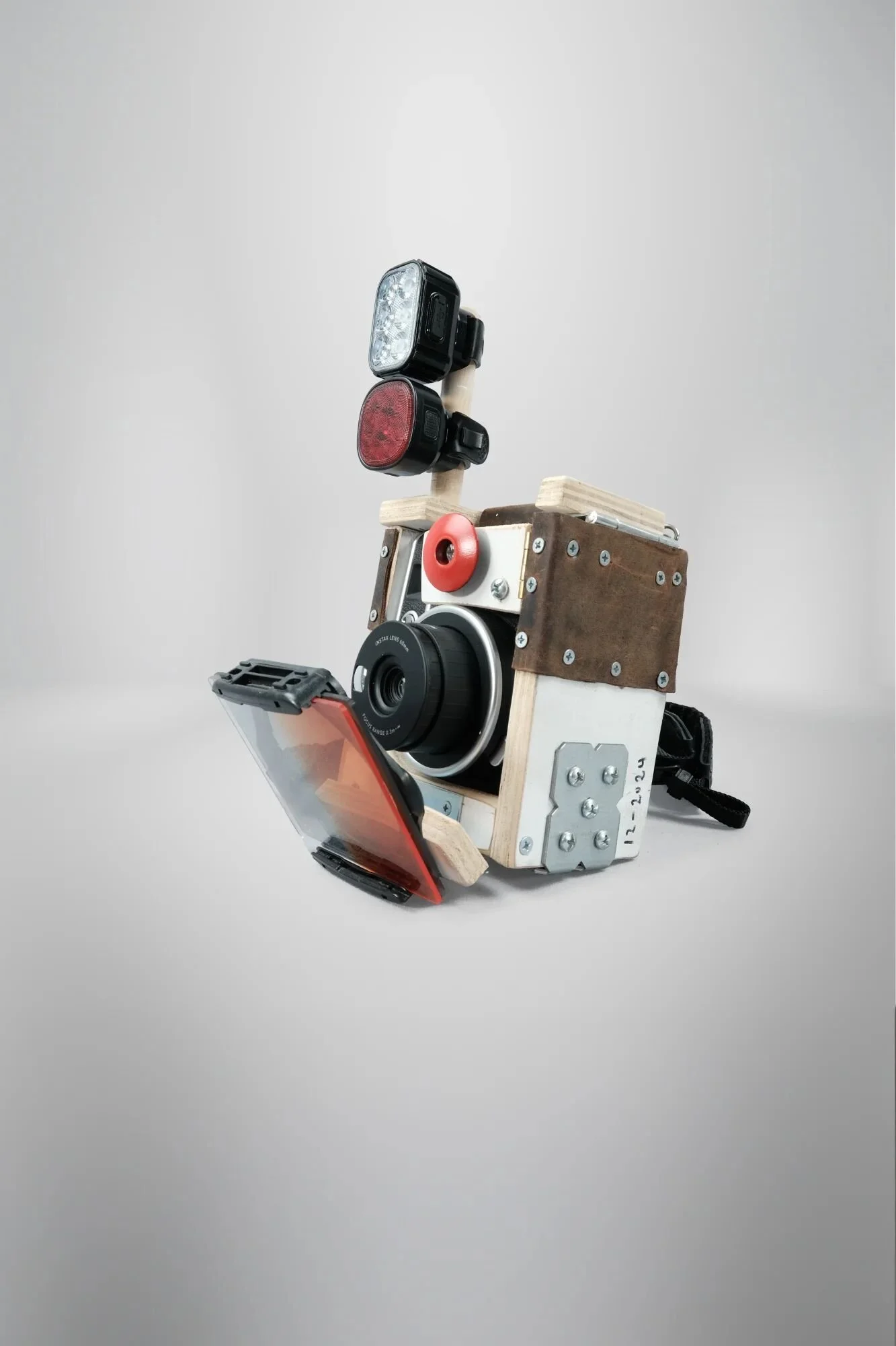 Bricolage Polaroid camera built around salvaged Fujifilm Instax Mini 40 body. Dual LED bike lights mounted on wooden rods, plywood housing with toggle clamp, ND filter mechanism, all hardware visible. The visible strangeness functions as provocation—