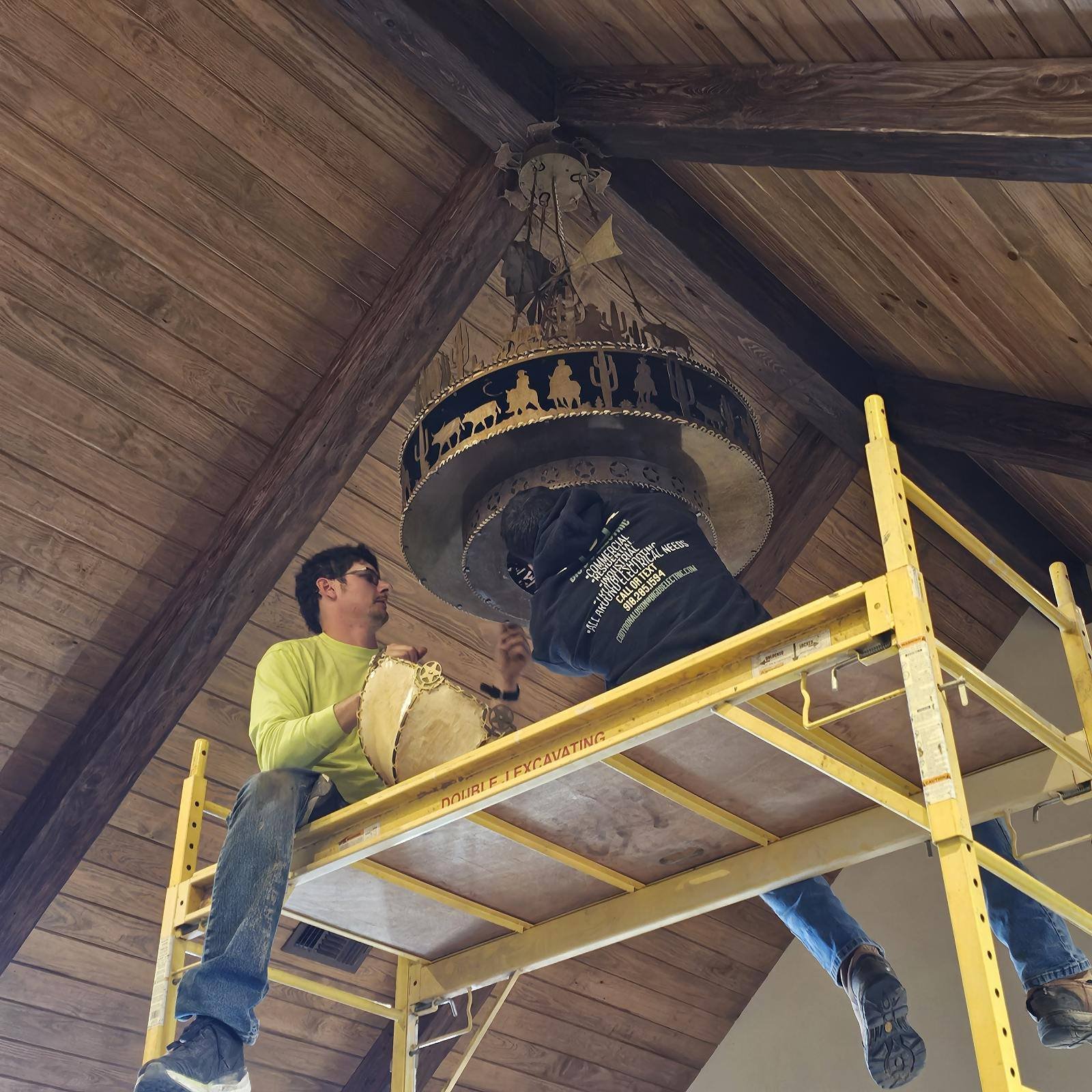 Two guys from Big Ds Electric installing a decorative light fixture with bronze cowboys herding cows. The shape is circular and has the cowboy design around the rim. This is on a wooden arched roof.