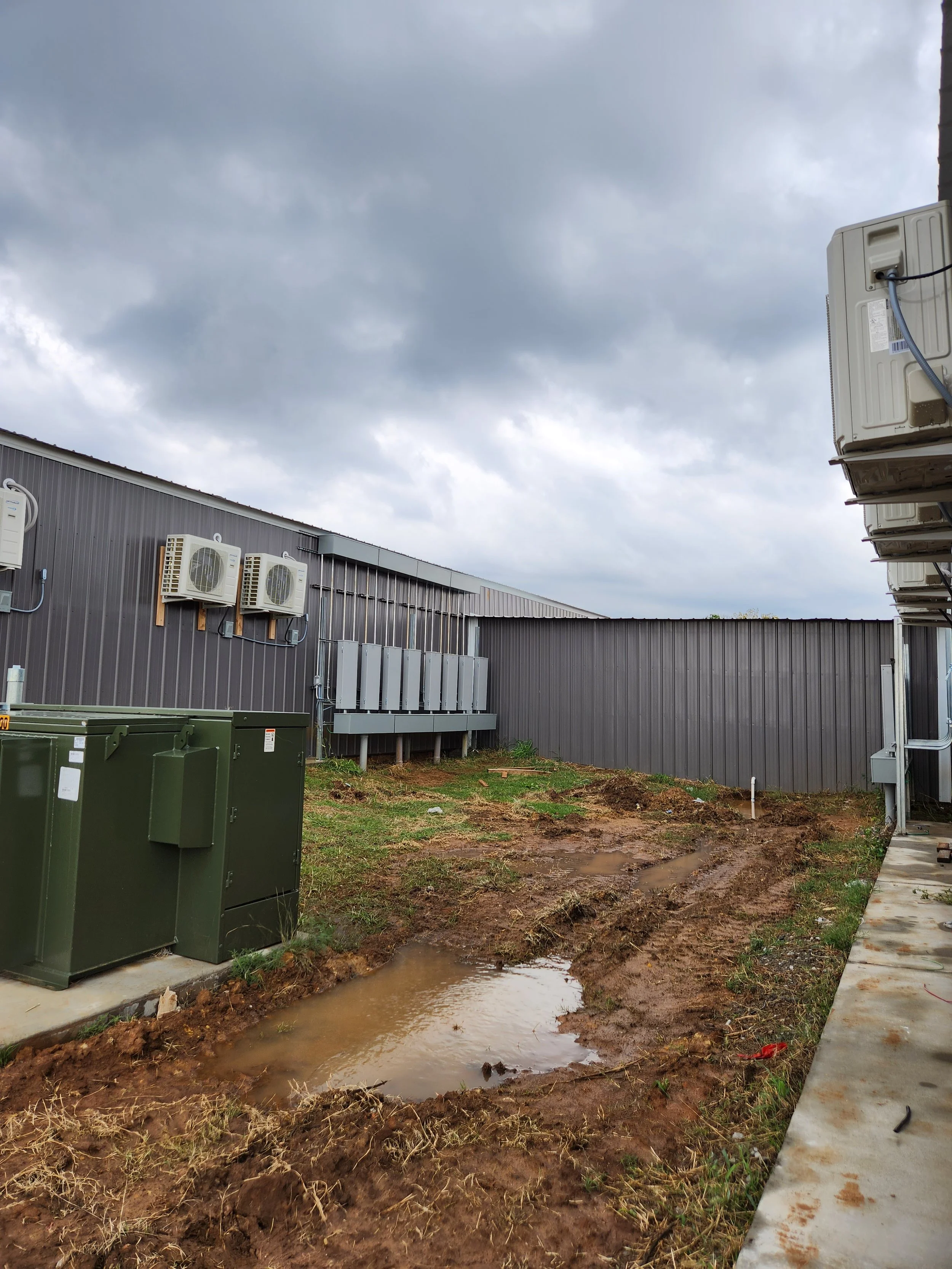 Construction site with muddy ground and puddles near an industrial building with HVAC systems, electrical panels, and ventilation units under an overcast sky.