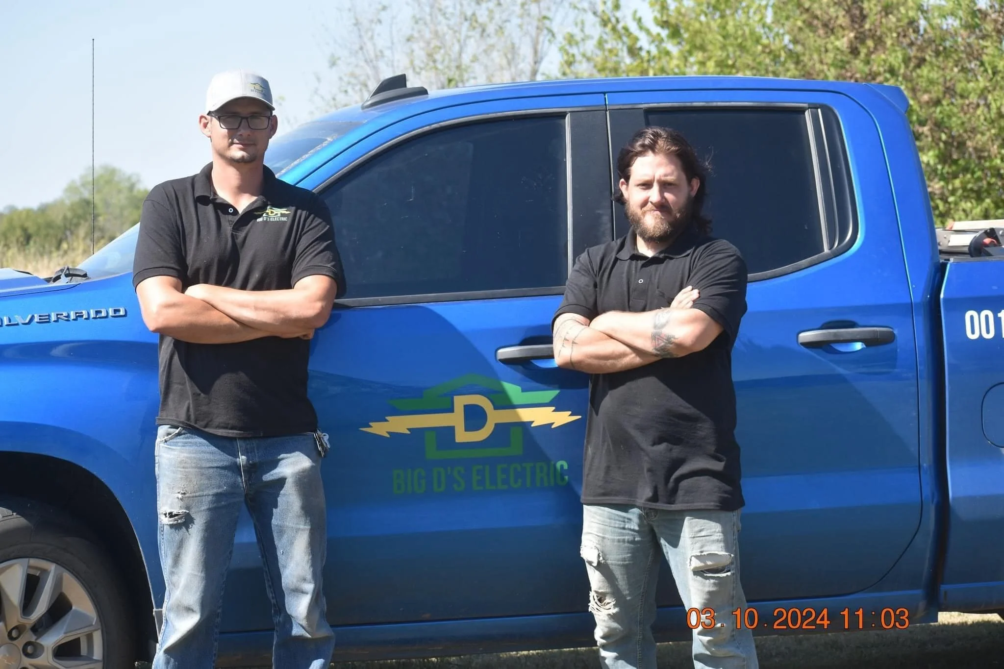 Two men standing with arms crossed in front of a blue pickup truck labeled 'Big D's Electric' on a grassy outdoor area.