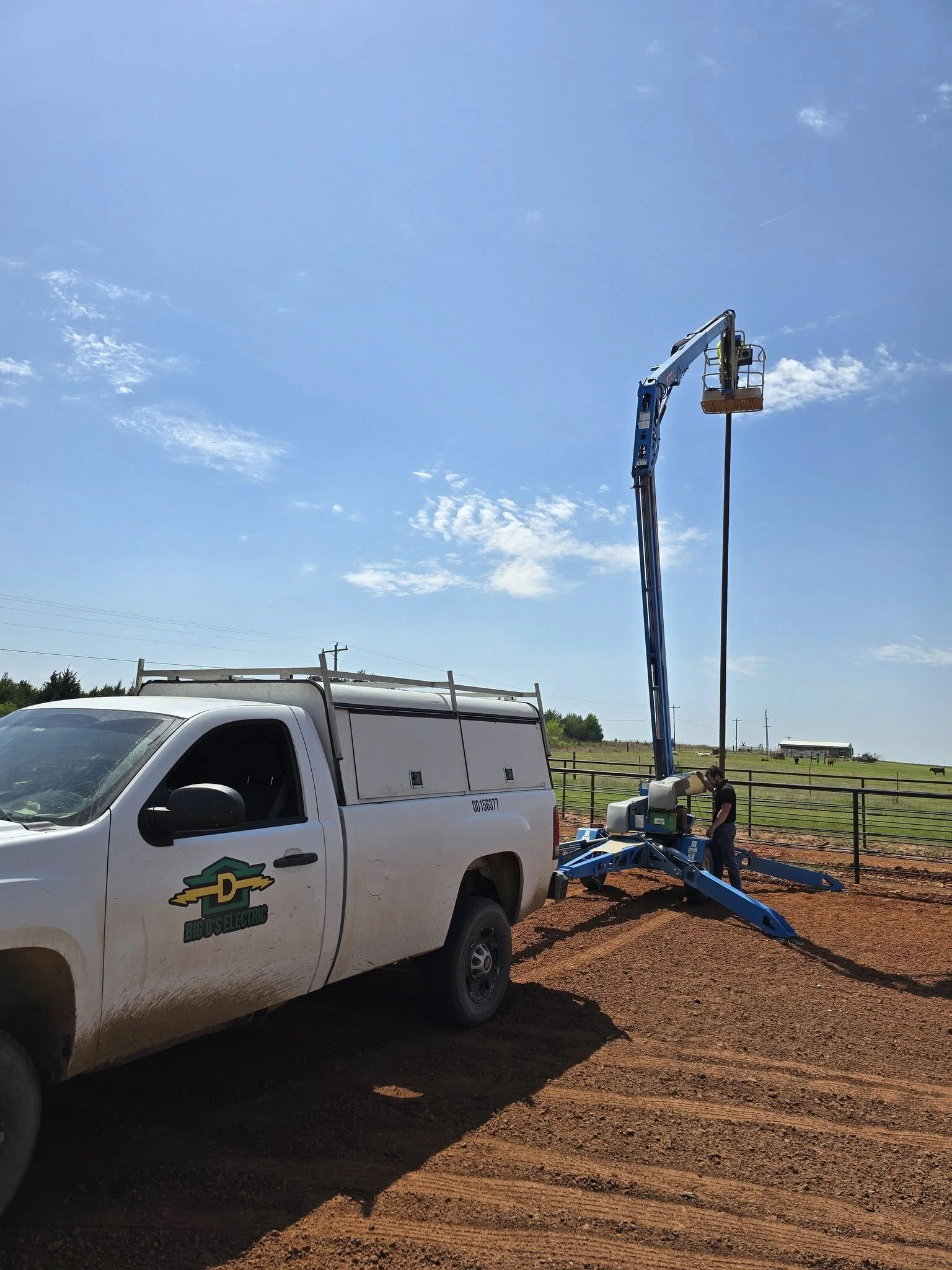 A white utility truck with logo and text 'Big D's Electric' parked on a dirt lot with a man working next to a tall blue cherry picker lift, which is extended high into the sky with a worker in the basket. The background shows open fields, a fence, and a clear blue sky with a few clouds.