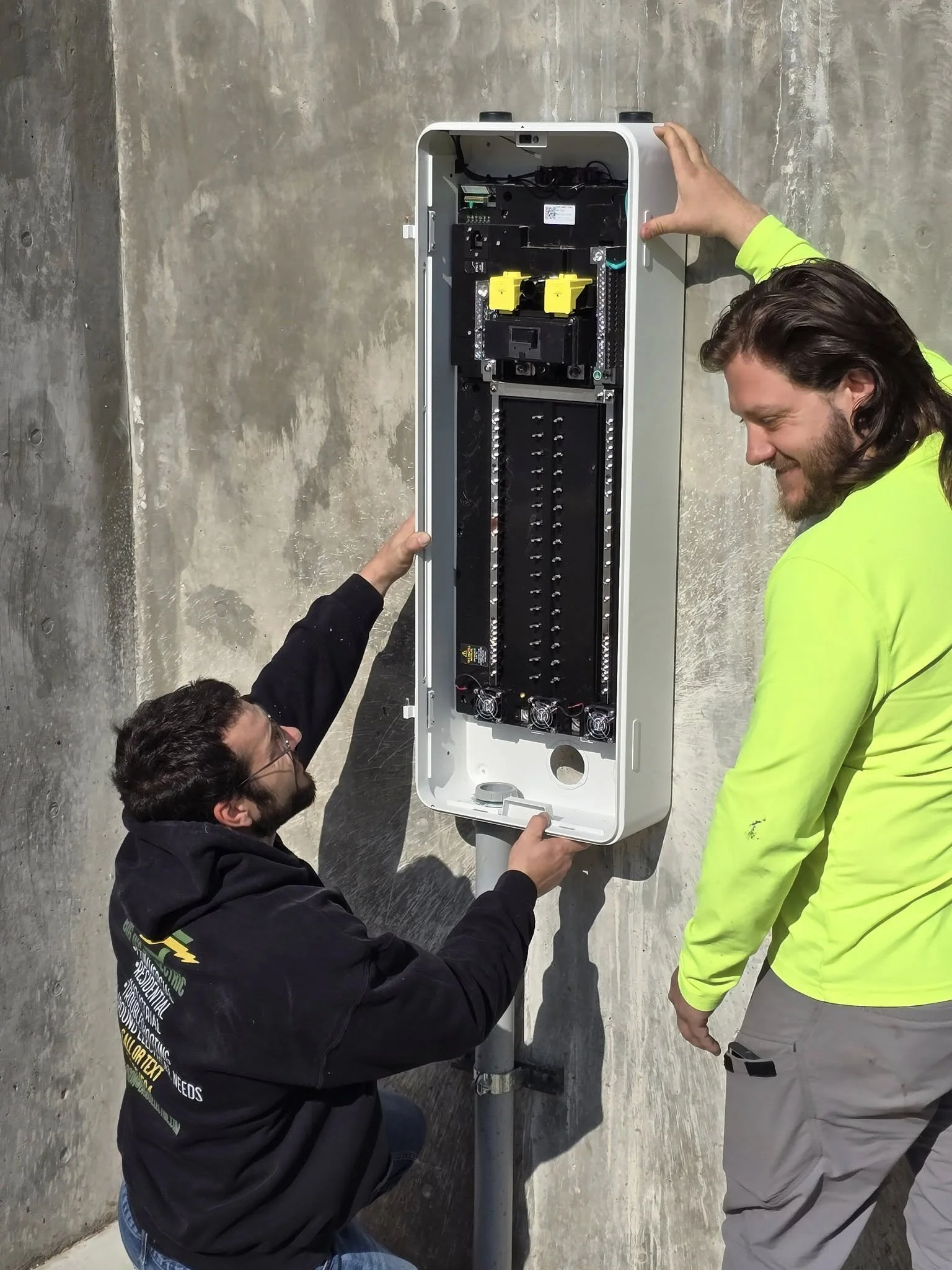 Two men installing or inspecting an outdoor electrical or fiber optic box mounted on a pole against a concrete wall.