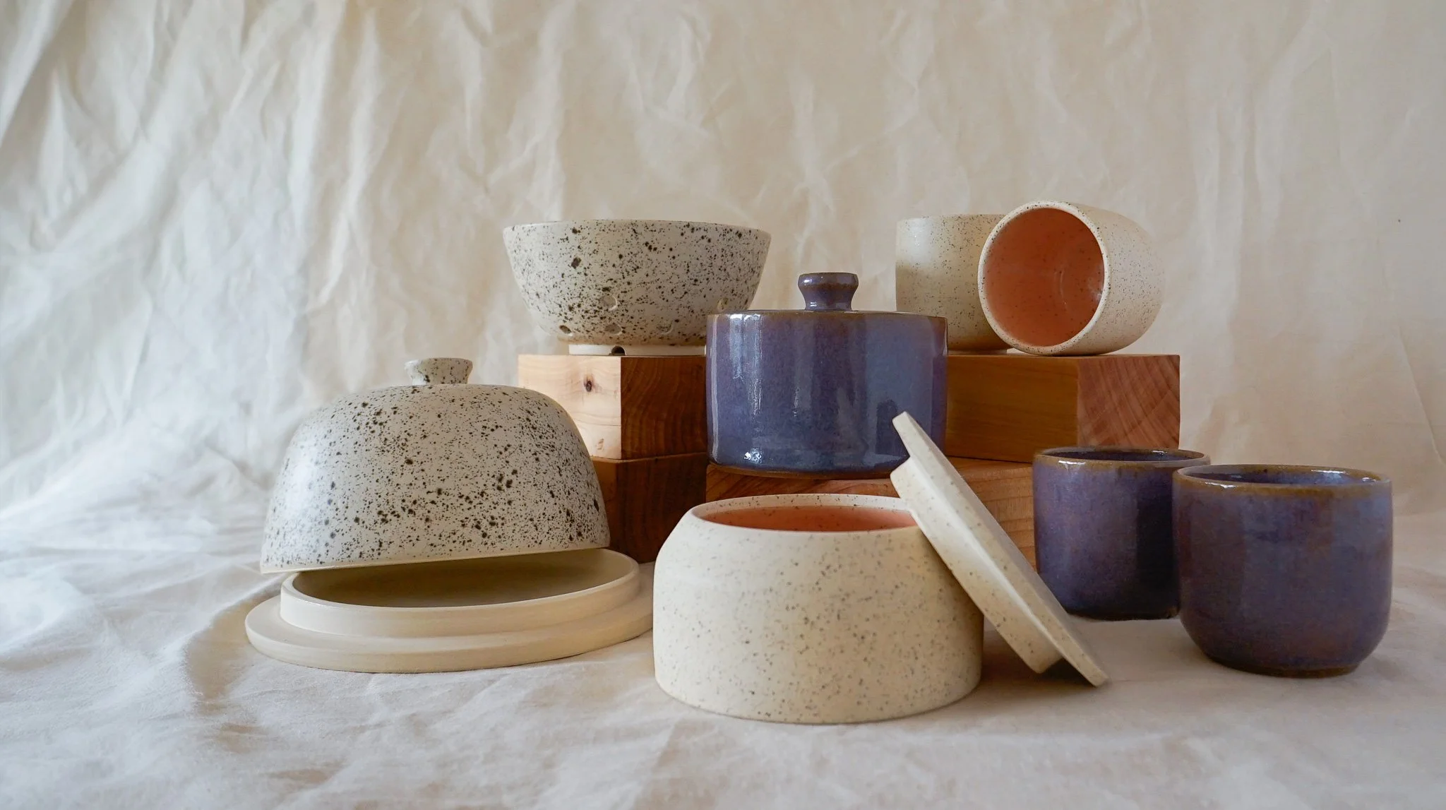Collection of ceramic bowls and cups in neutral tones, some with speckled patterns, arranged on wooden blocks and a draped fabric background.