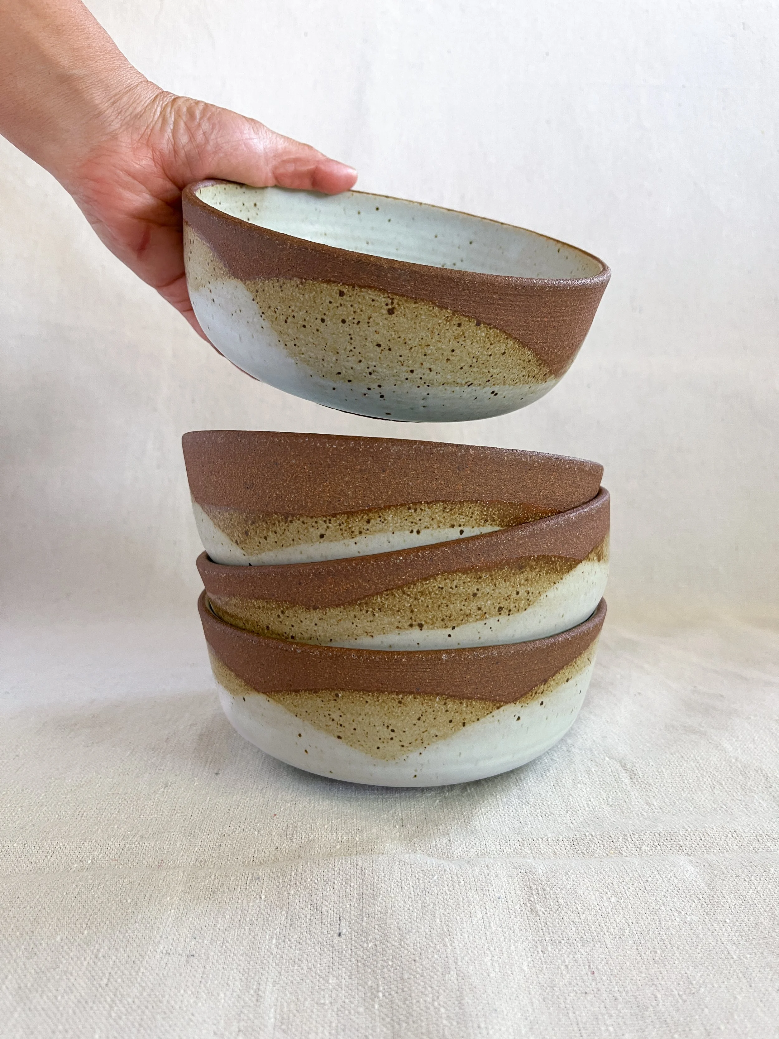 A person holding a ceramic bowl above three stacked similar bowls on a beige fabric surface.