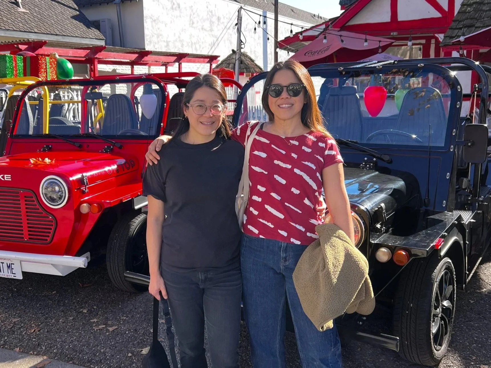 Two women standing next to each other with their arms around each other's shoulders, smiling. They are in front of a red and black mini Jeep vehicle with a clear windshield and decorative lights inside. The background features a Christmas-themed outdoor setup with red and white striped umbrellas, string lights, and a snowy landscape.