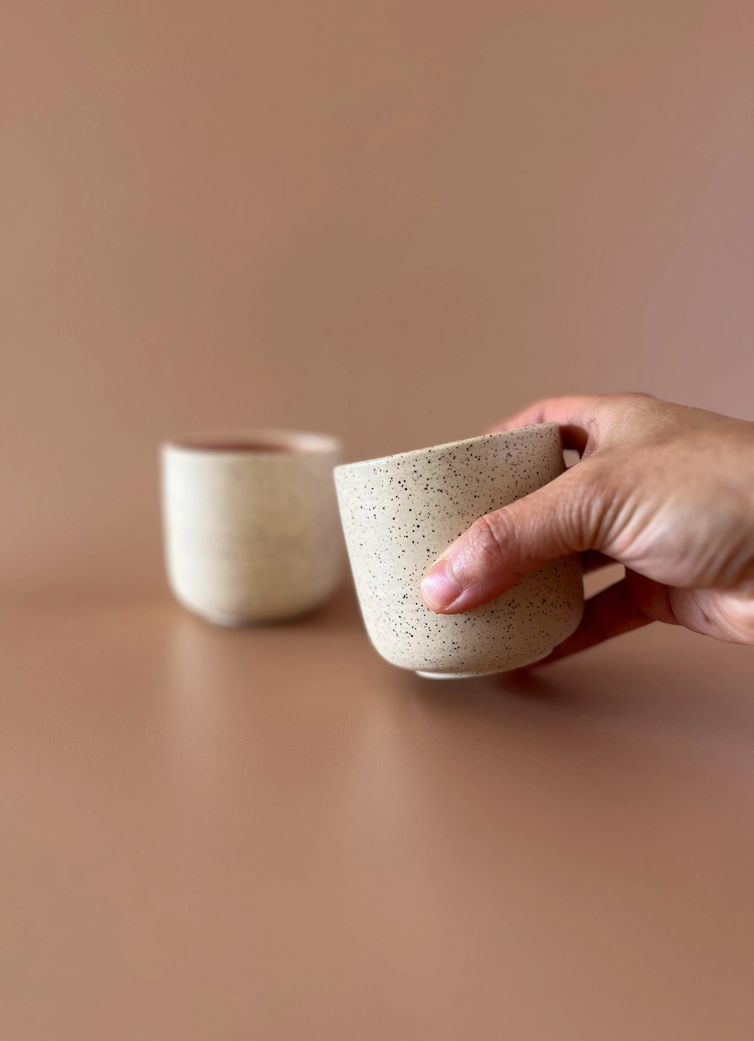 Two beige speckled ceramic cups on a brown surface, one held in hand, the other slightly blurred in the background.
