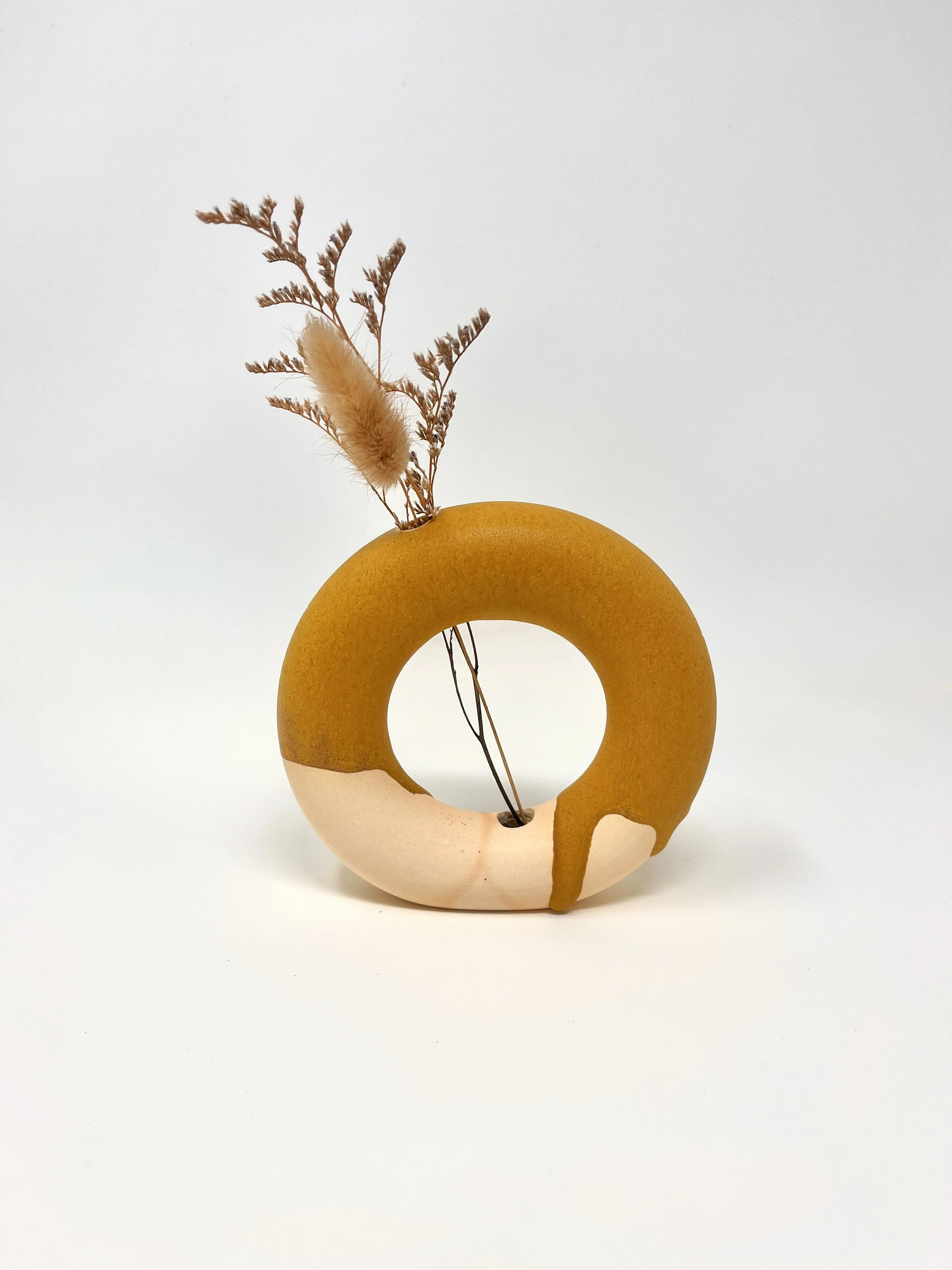 Decorative ceramic vase with a circular opening, partially glazed in white and mustard yellow, holding dried grass and a fluffy, cylindrical flower stem, set against a plain white background.