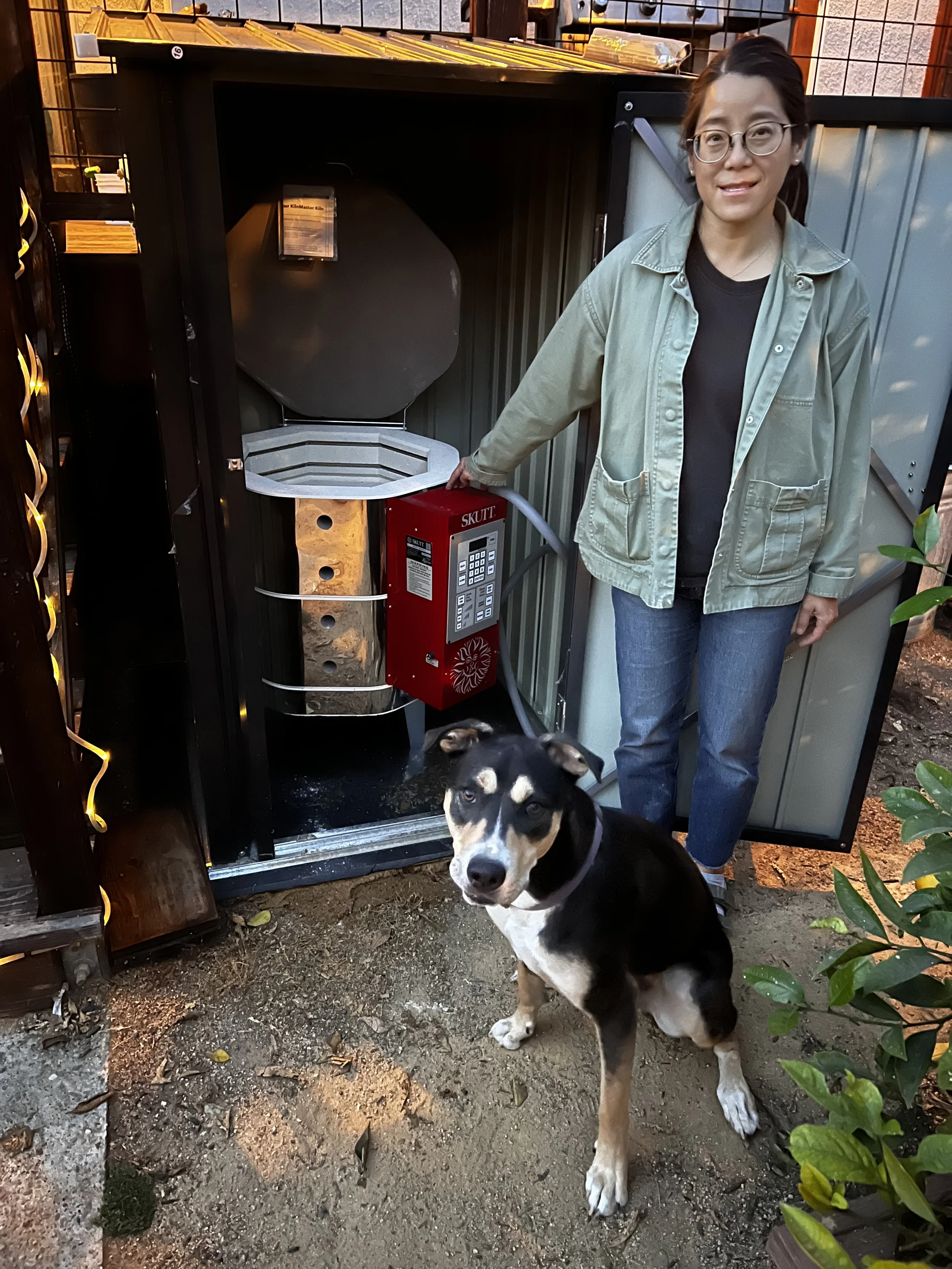 A woman standing next to an outdoor shed with a kiln inside, a black and tan dog sits in front of her, outdoors with plants around.