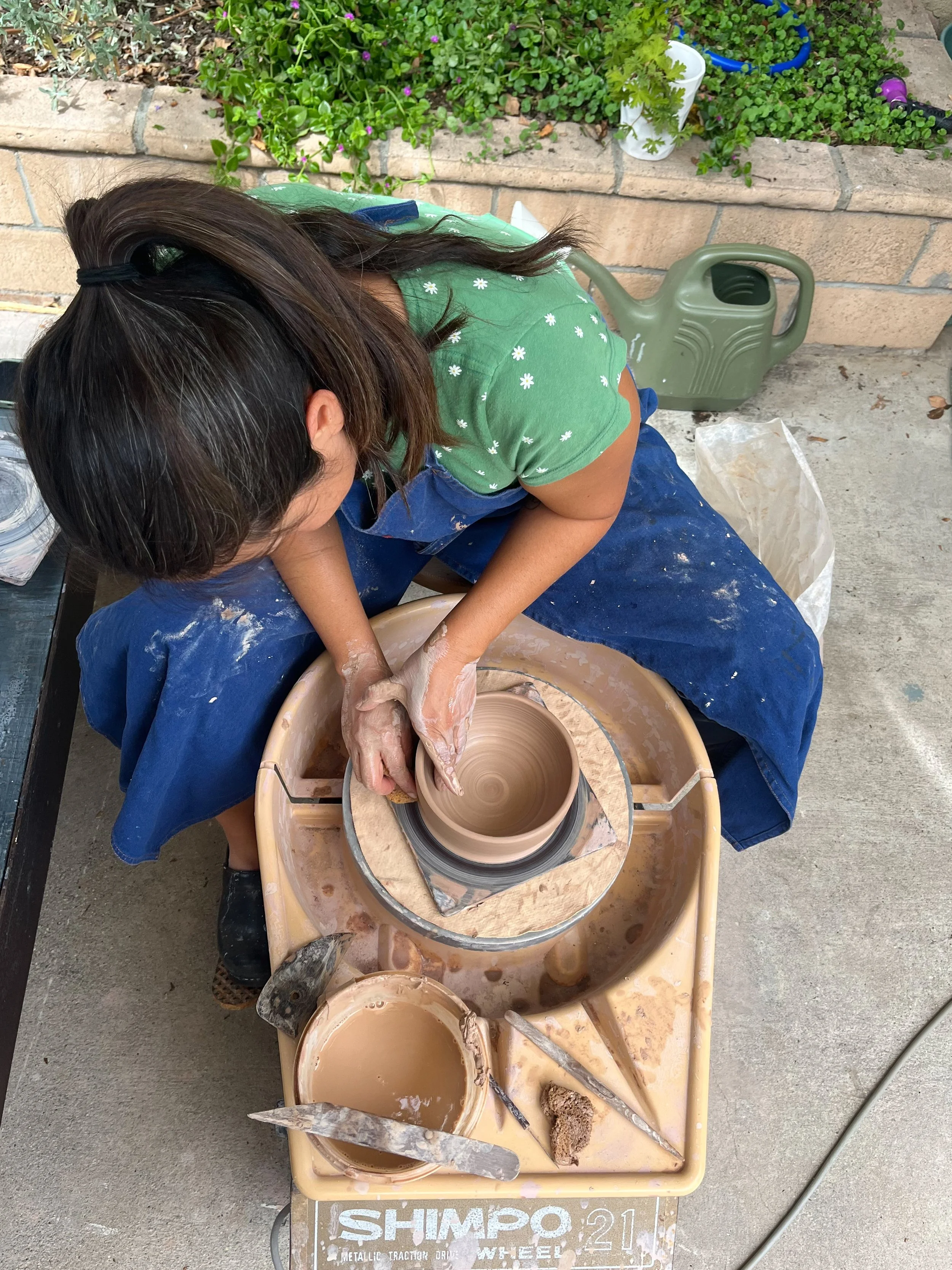 A woman working on a pottery wheel, shaping clay into a vessel outside. She is wearing a green shirt with white flowers and blue pants, with various tools and wet clay around her.