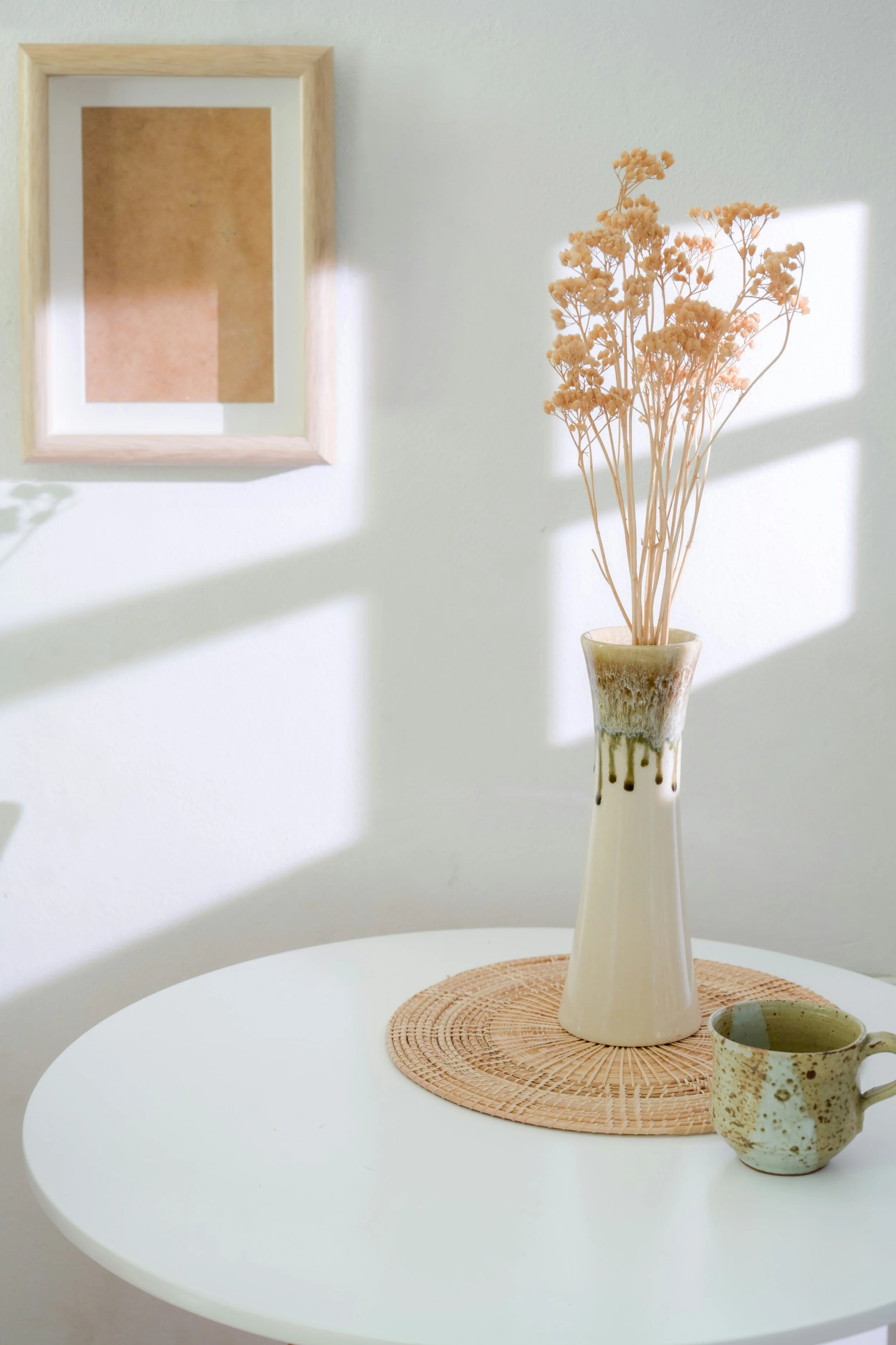 A white table with a round woven placemat, a tall ceramic vase with dried flowers, and a speckled ceramic mug. Sunlight casts shadows on the white wall behind.