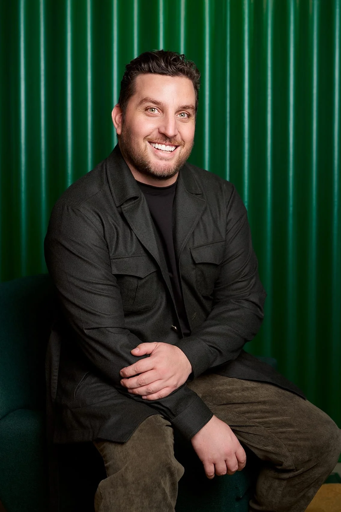 A smiling man with dark hair and beard, dressed in black jacket and brown pants, sitting on a green chair in front of a green curtain.