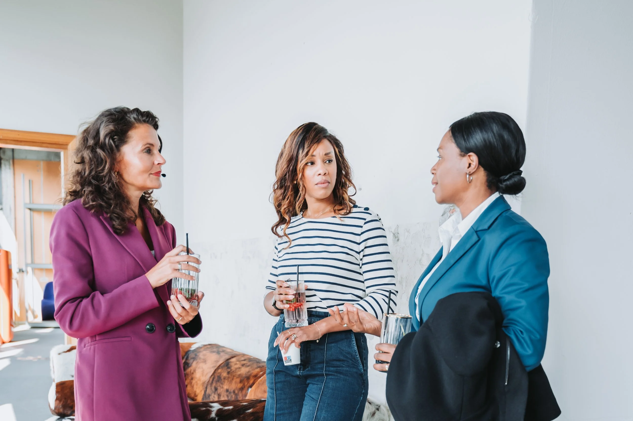 Three women having a conversation indoors, two holding glasses with drinks, one woman gesturing with her hand.