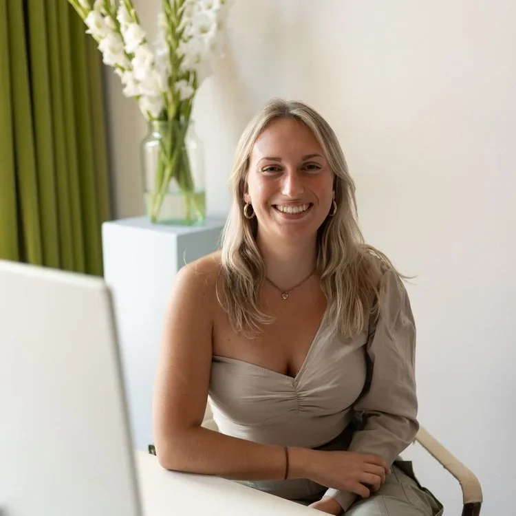A smiling woman with blonde hair wearing a beige off-shoulder top sitting at a desk with a computer, in a room with white walls, green curtains, and a vase of white flowers in the background.