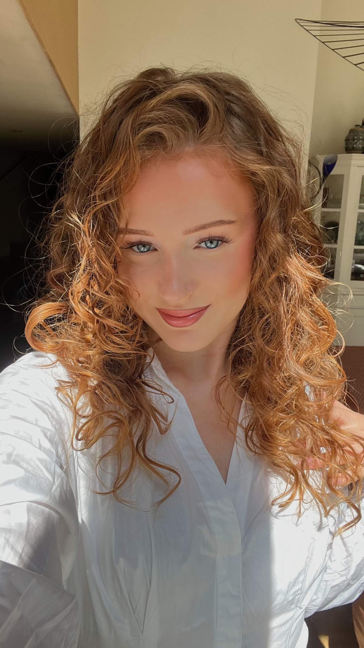 A woman with curly red hair and blue eyes taking a selfie indoors, wearing a white shirt.