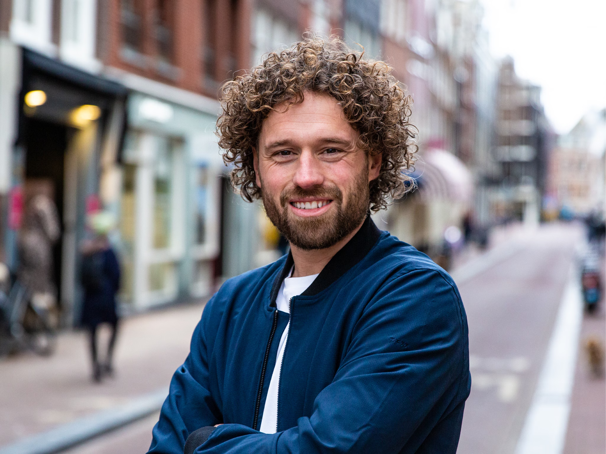 A smiling man with curly hair and a beard wearing a blue jacket standing on a city street.