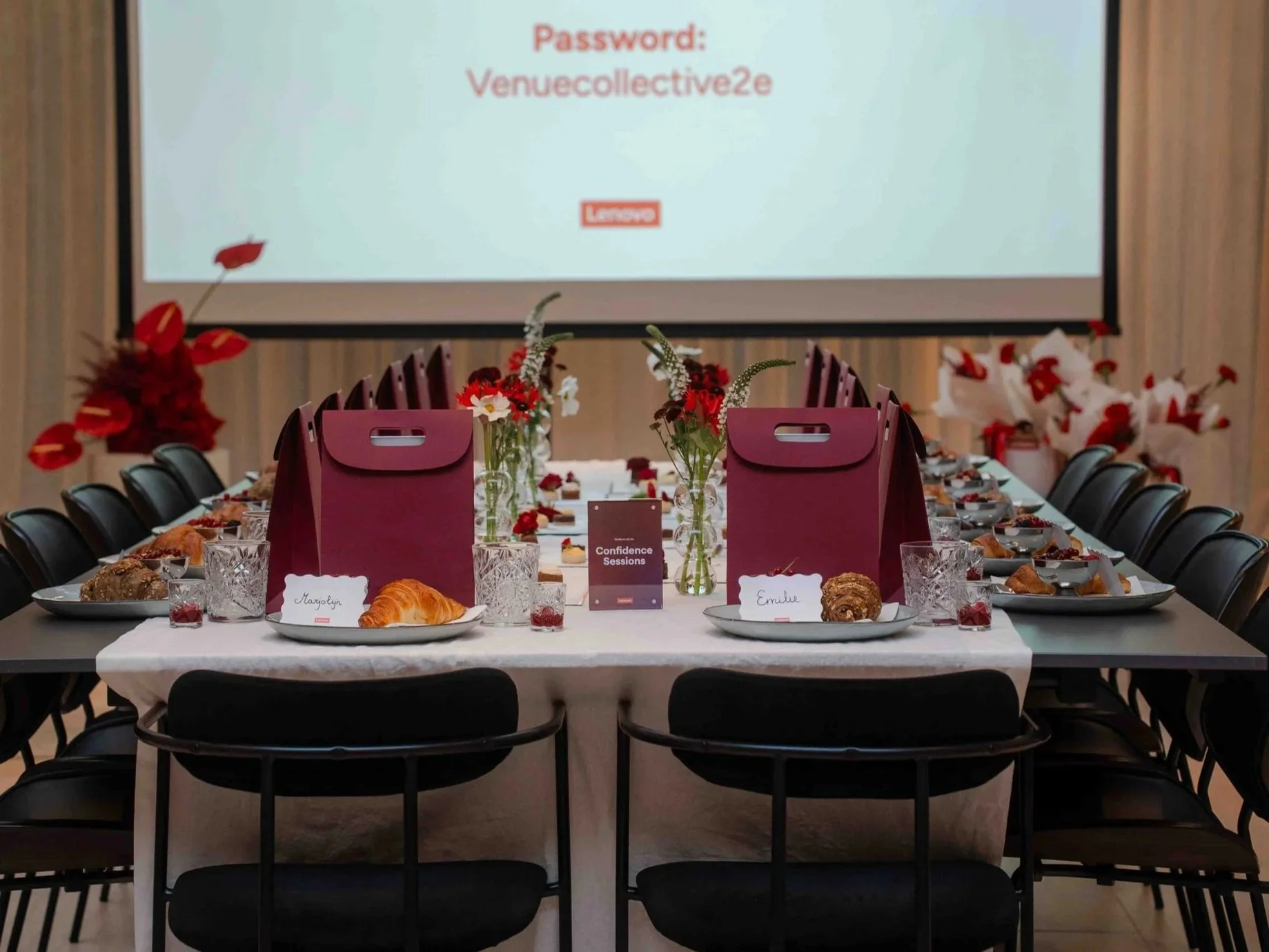 A conference room decorated for a meeting with a long table set with pastries, flowers, and burgundy gift bags, and a large screen displaying a password and the words 'Confidence Sessions'.