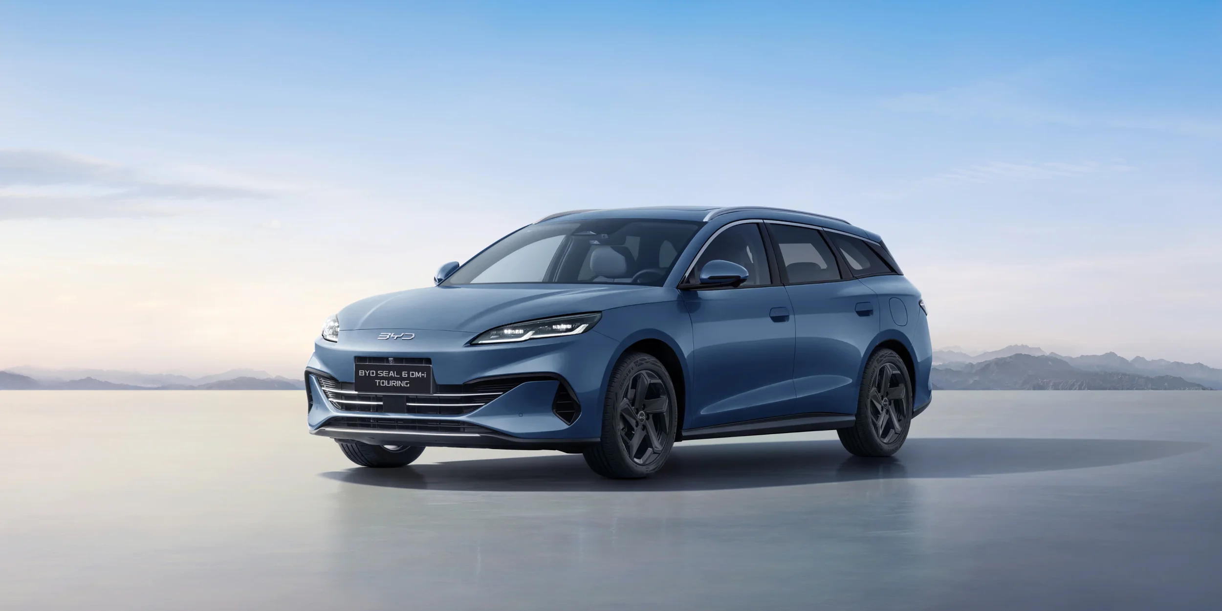 A blue BYD Seal 6 DM-i Touring electric vehicle parked on a flat surface with mountains and a cloudy sky in the background.