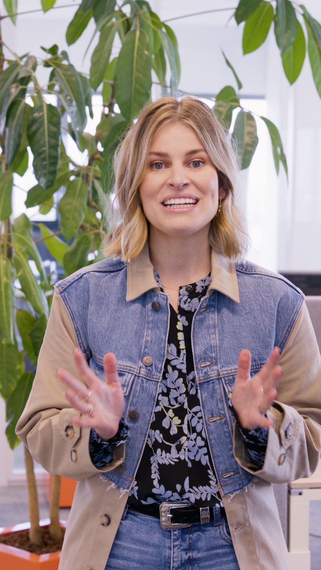 A young woman with shoulder-length blonde hair, wearing a denim and beige coat, and a black floral shirt, is smiling and gesturing with her hands in an indoor setting with large green plants.