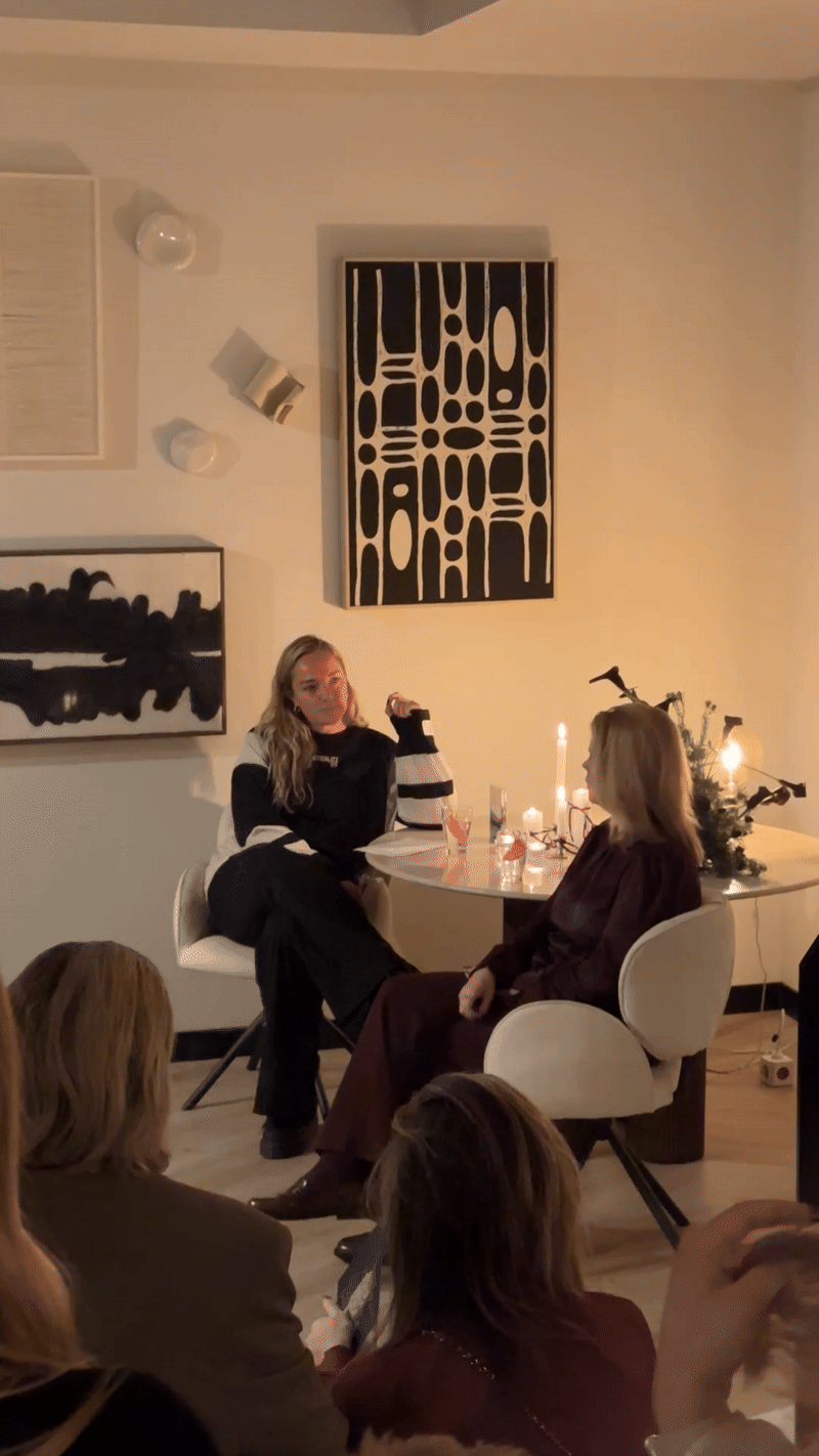 Two women sitting at a round table filled with candles and drinks, having a conversation in a warmly lit indoor setting with modern art on the wall and a small Christmas tree in the background.