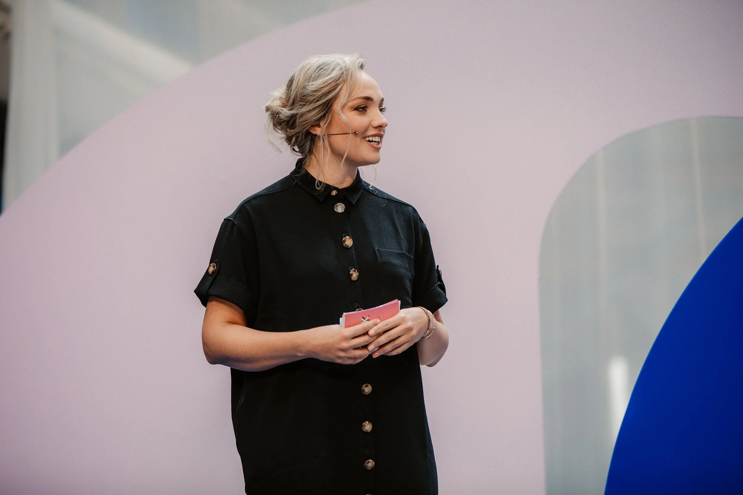 A woman with platinum blonde hair styled in loose waves, smiling, wearing a black button-up shirt, standing in front of a pink and blue backdrop.