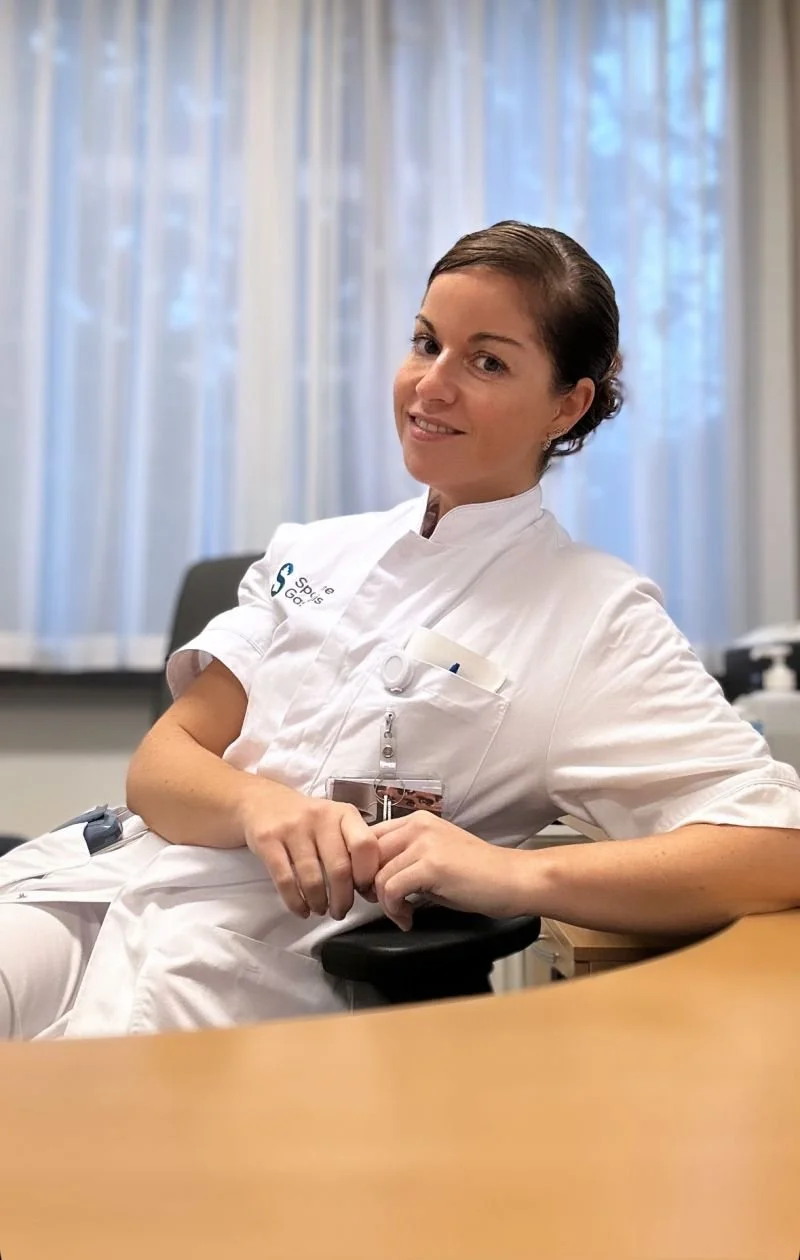 A woman wearing a white medical uniform sitting in an office or conference room, smiling at the camera, with a window and light curtains in the background.