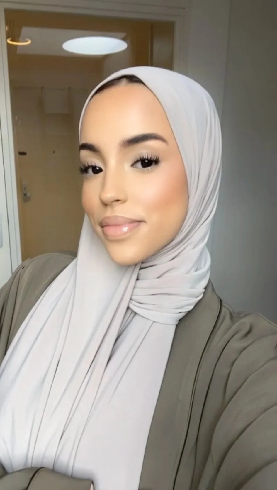 A woman wearing a light-colored hijab and beige jacket taking a selfie in front of a mirror.