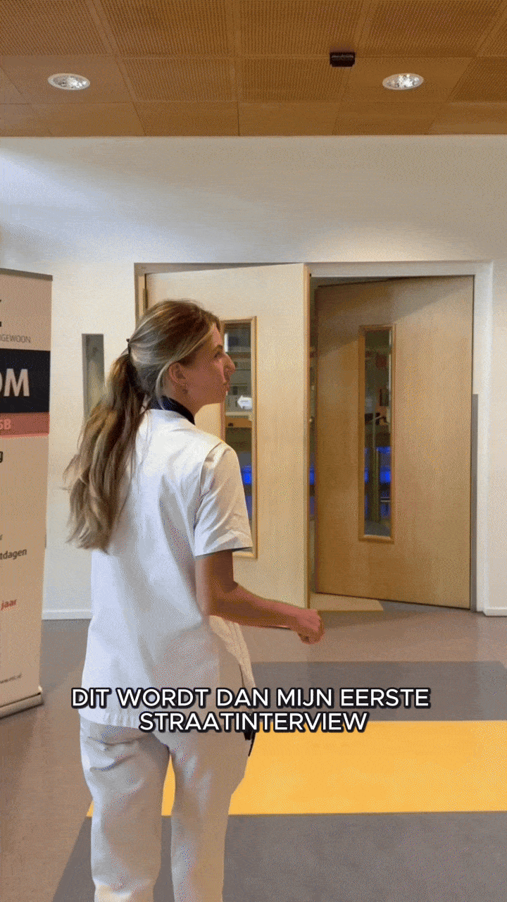 A woman dressed in medical attire, standing inside a building, possibly a hospital or clinic, with a wooden ceiling and doors behind her. There is a yellow mat on the floor and a sign on the left side of the image. The woman appears to be giving her first street interview, as indicated by the Dutch caption.