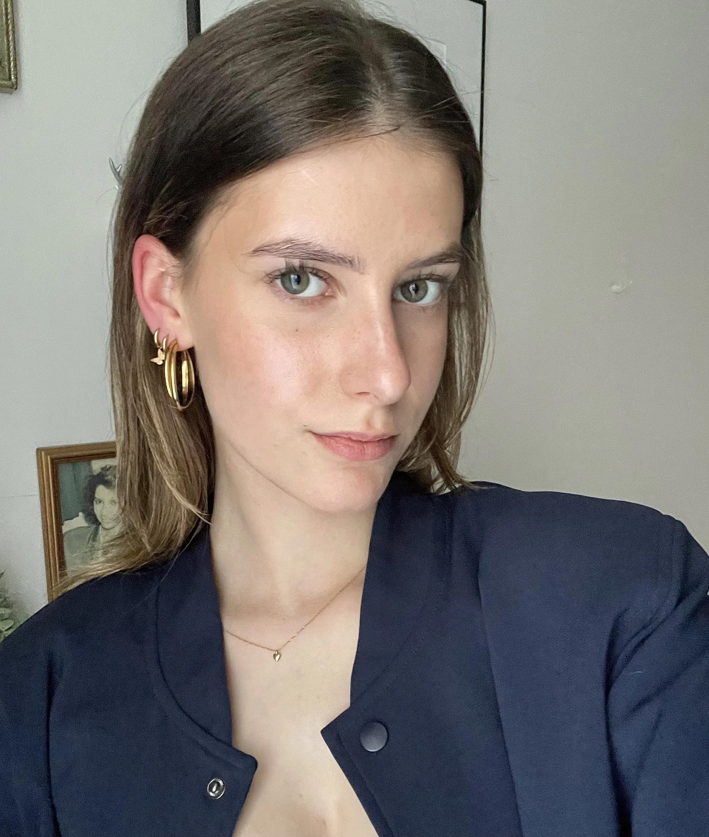 Close-up selfie of a young woman with shoulder-length brown hair, blue eyes, gold hoop earrings, a delicate necklace, and a dark blue shirt, standing in front of a wall with framed photographs.