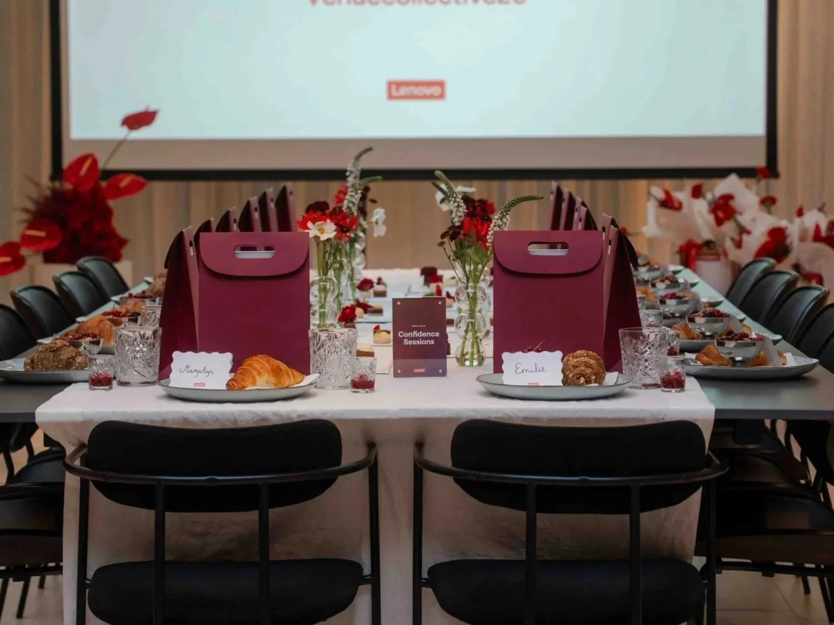 A decorated conference event table set with plates of croissants, glasses, flowers, and gift bags, with a large screen in the background and white walls.