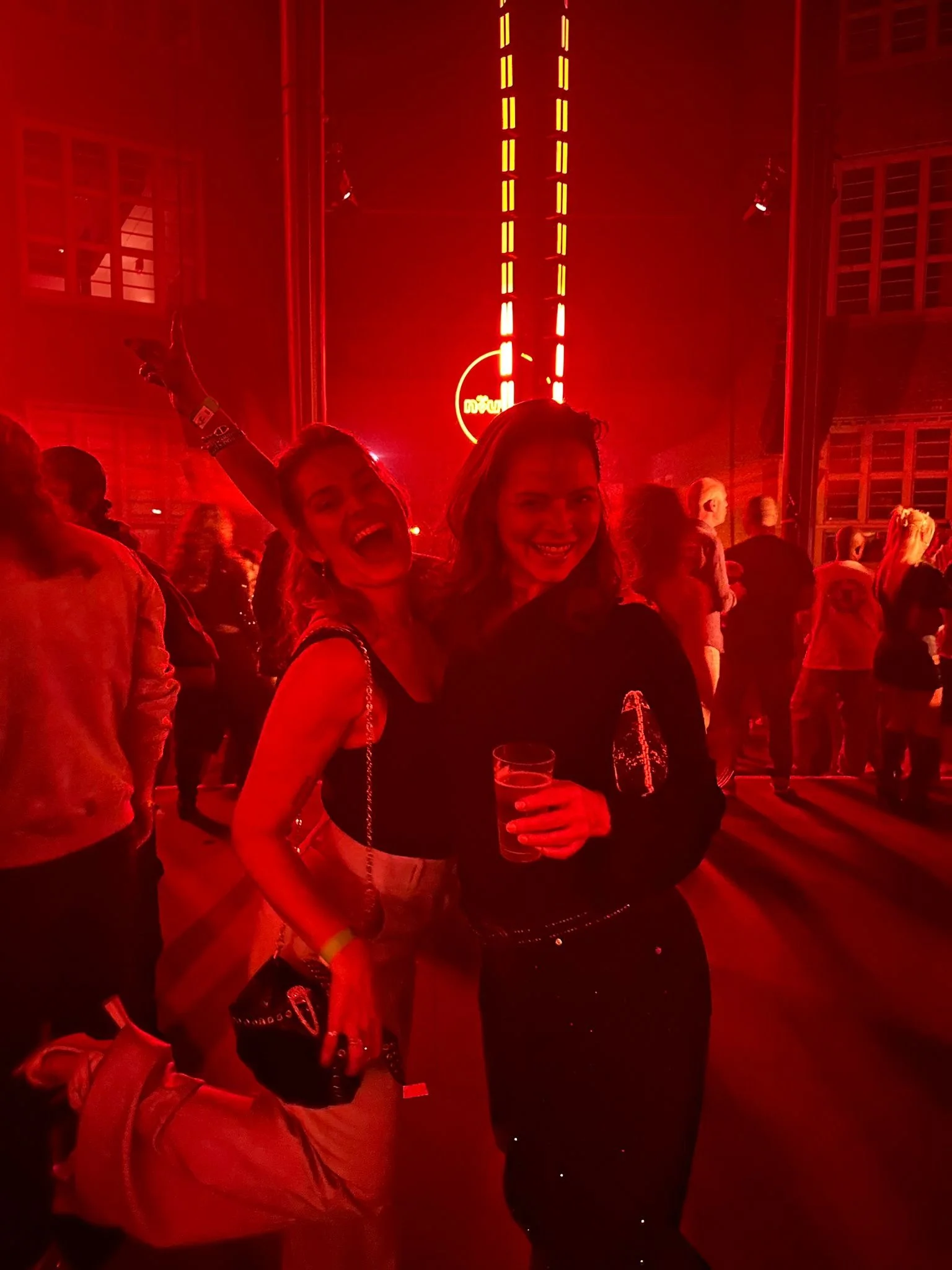 Two women smiling and posing for the camera in a crowded indoor event, with red lighting and neon signs in the background. One woman is holding a drink.