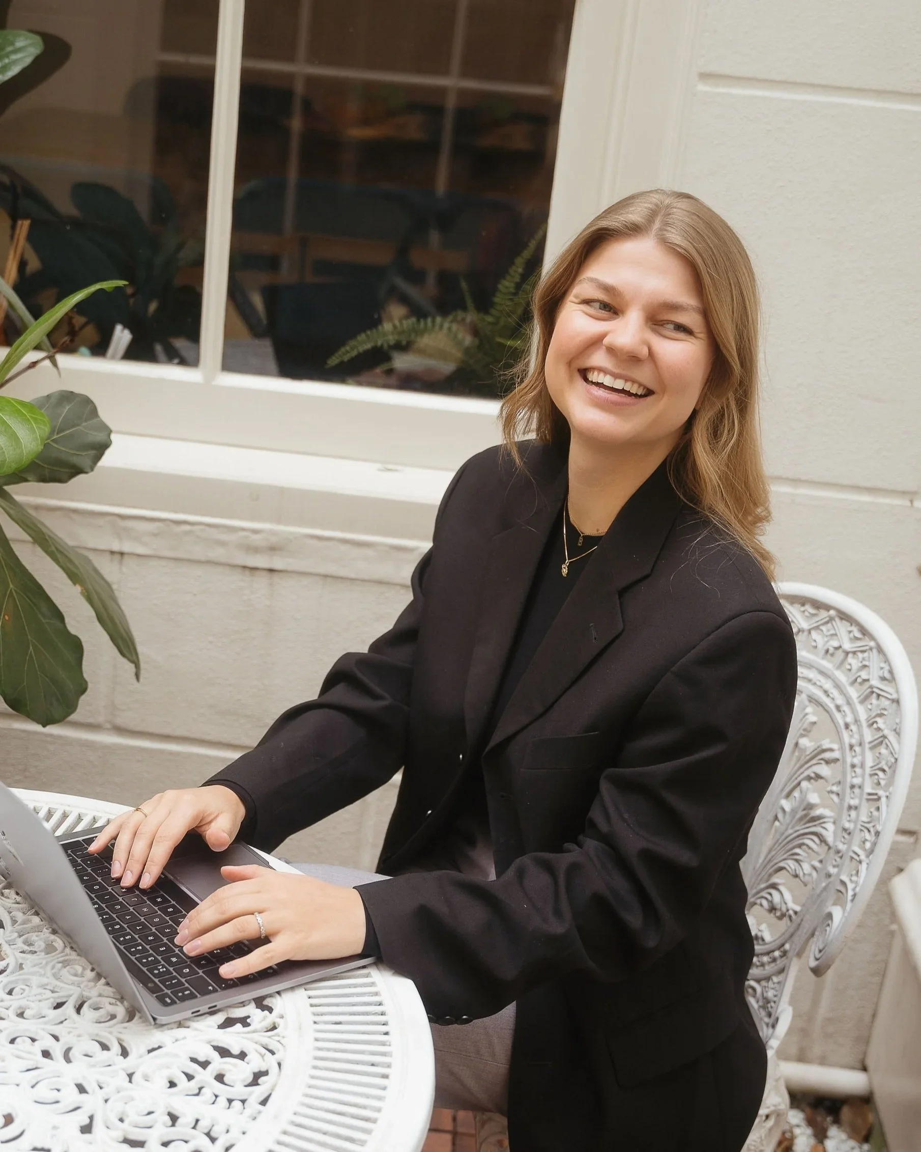 A smiling woman in a black blazer working on a laptop at an ornate white table indoors, with a window and potted plants in the background.