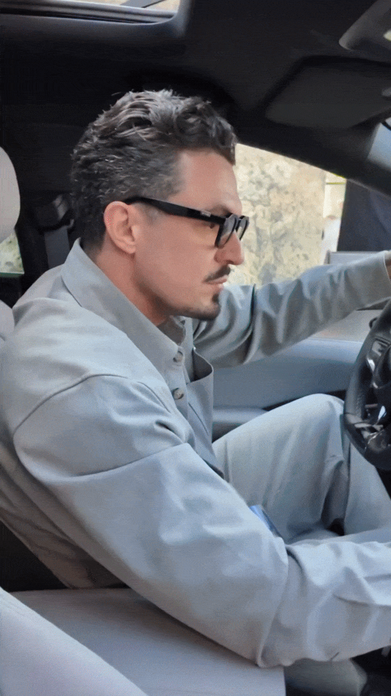 A man with dark, slicked-back hair and glasses driving a vehicle, wearing a light gray shirt, with a focused expression.