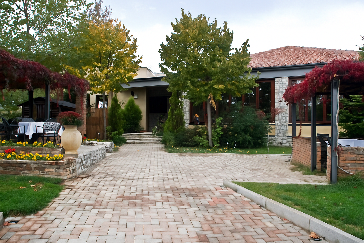 Η πέτρινη αυλή στο Φαγοτόπι με δέντρα, θάμνους και πλακόστρωτο μονοπάτι. Θέα προς το εστιατόριο με κεραμοσκεπή και μεγάλες τζαμαρίες.