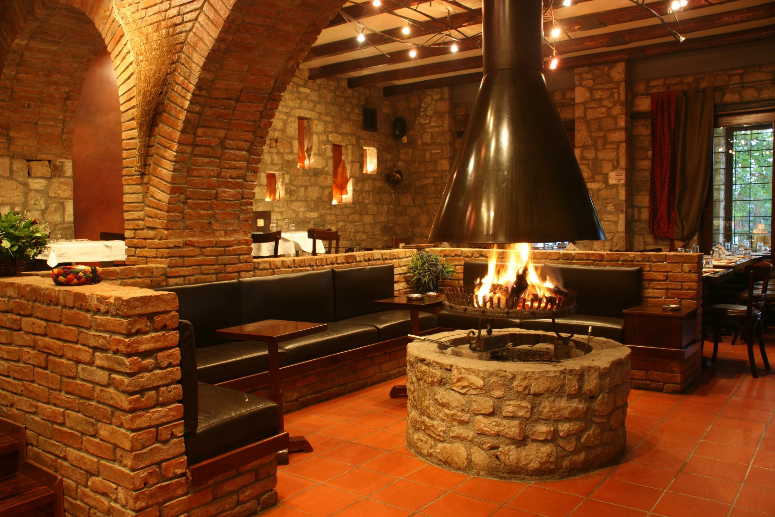 Εσωτερικός χώρος στο Φαγοτόπι με τούβλινη καμάρα, κεντρικό τζάκι και τραπέζια στρωμένα με λευκά τραπεζομάντηλα. Ζεστή ατμόσφαιρα με κόκκινες κουρτίνες και παραδοσιακή διακόσμηση.
