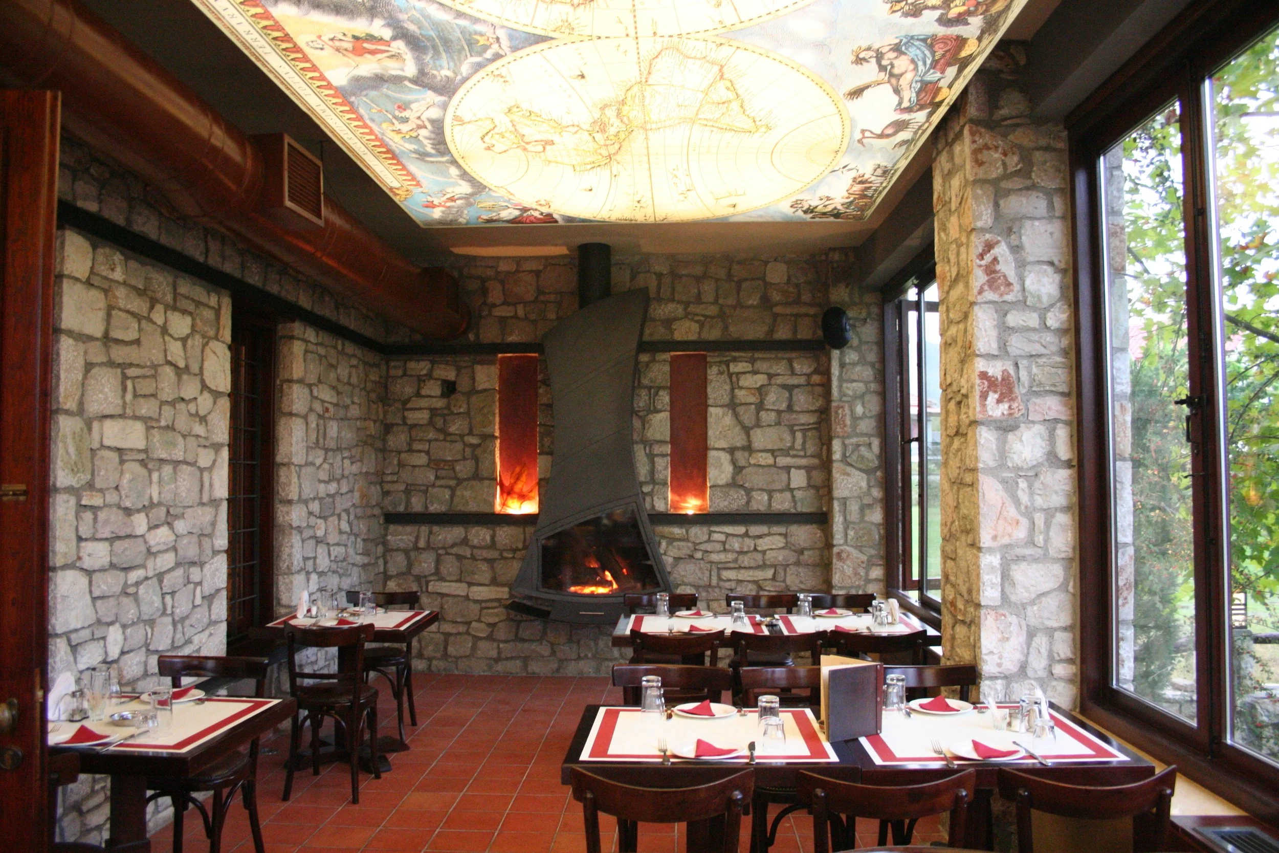 Ζεστός εσωτερικός χώρος στο Φαγοτόπι με πέτρινους τοίχους, ξύλινα έπιπλα, τζάκι και μεγάλες τζαμαρίες με θέα στο πράσινο.
