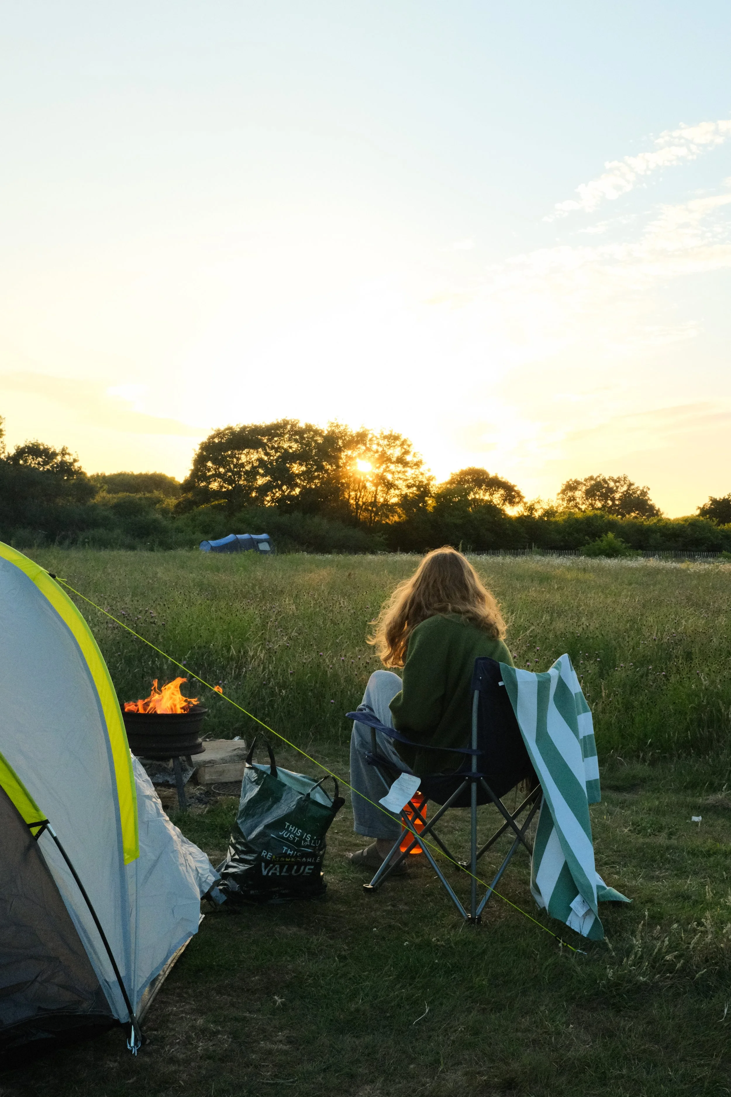 A girl sat around a campfire during sunset in a wild flower meadow
