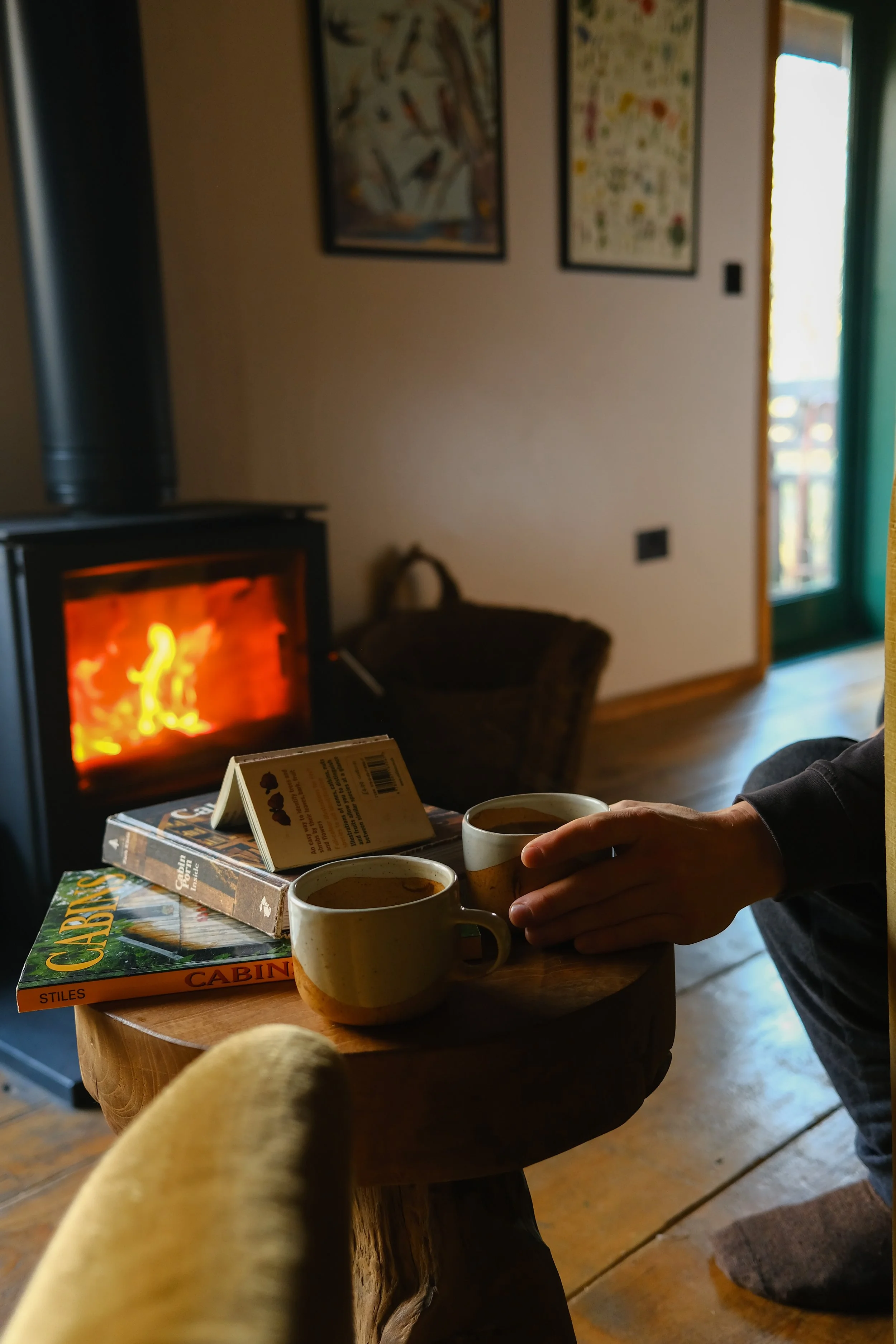 A cosy cabin where coffee cups sit next to books on a table with a log burner glowing