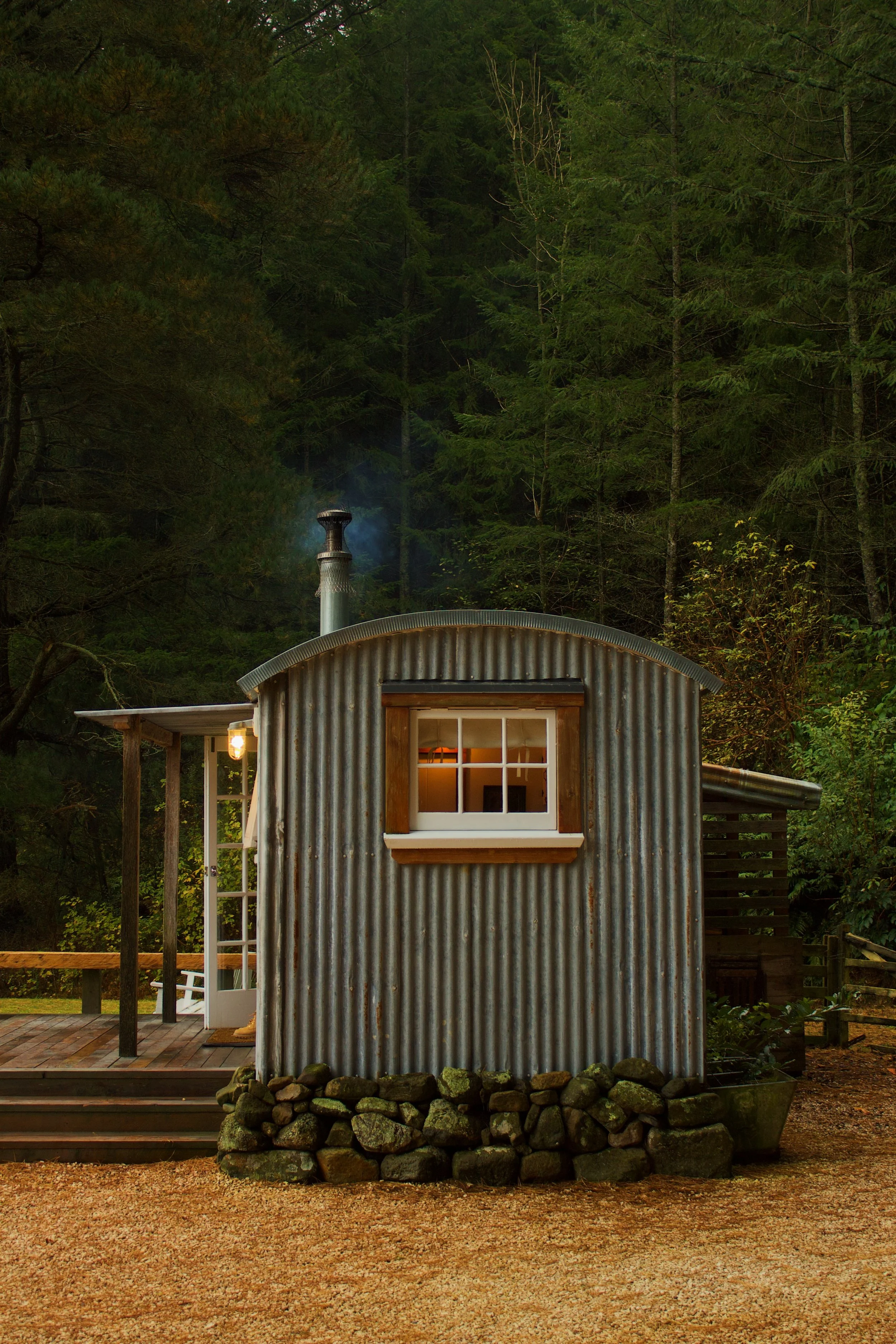 A small shepherds hut in New Zealand surrounded by trees, captured from the outside with glowing windows and smoke coming out the chimney