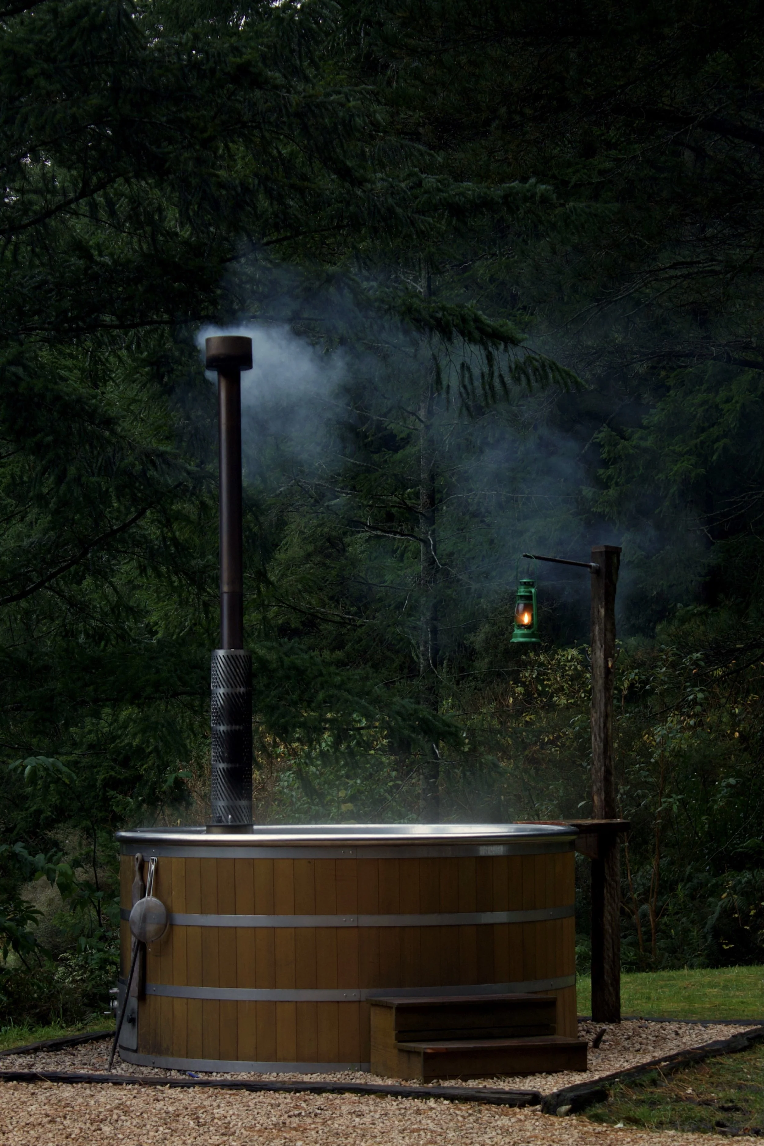 A wood fired hot tub steaming with a glowing lantern during evening against dark green trees