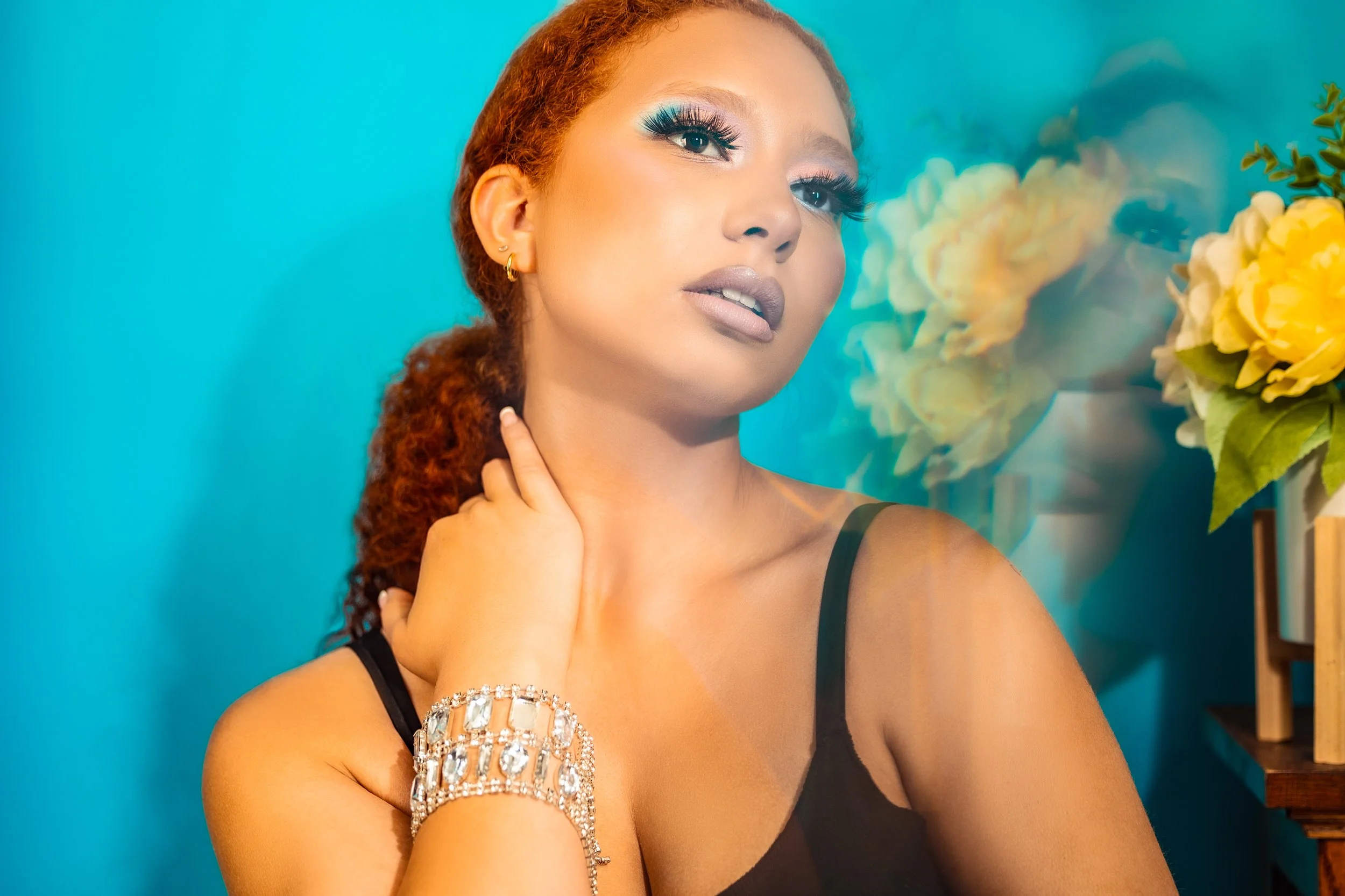 A woman with red curly hair, wearing makeup with blue eyeshadow and long eyelashes, is posing in front of a turquoise background. She is wearing a black sleeveless top, gold earrings, and a sparkly bracelet. She is touching her neck with her left han